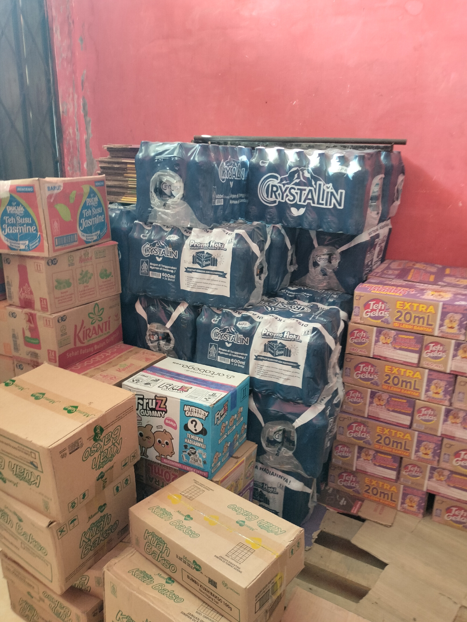 The image displays a storage area, likely within a retail or distribution facility in Langsa, Indonesia. The scene is filled with various stacked products, predominantly beverages and food items. Prominently featured are multipacks of bottled water labeled "CRYSTALIN" and boxes of tea. To the left, there are stacks of red and white boxes for "Pucuk Teh Susu Jasmine" and other boxes with green and red labels, some indicating "Kiranti". In the foreground and right side, there are stacks of boxes labeled "Teh Gelas EXTRA 20ml" and "FruZ Gummy". Several cardboard boxes with the label "Kuah Bakso" are also visible. The background shows a textured, reddish-pink wall with some vertical dark elements, possibly shelving or structural supports. The lighting is somewhat dim, casting shadows. There are no people present in the image, and the focus is entirely on the organized arrangement of goods. The overall impression is one of a well-stocked inventory ready for distribution or sale.