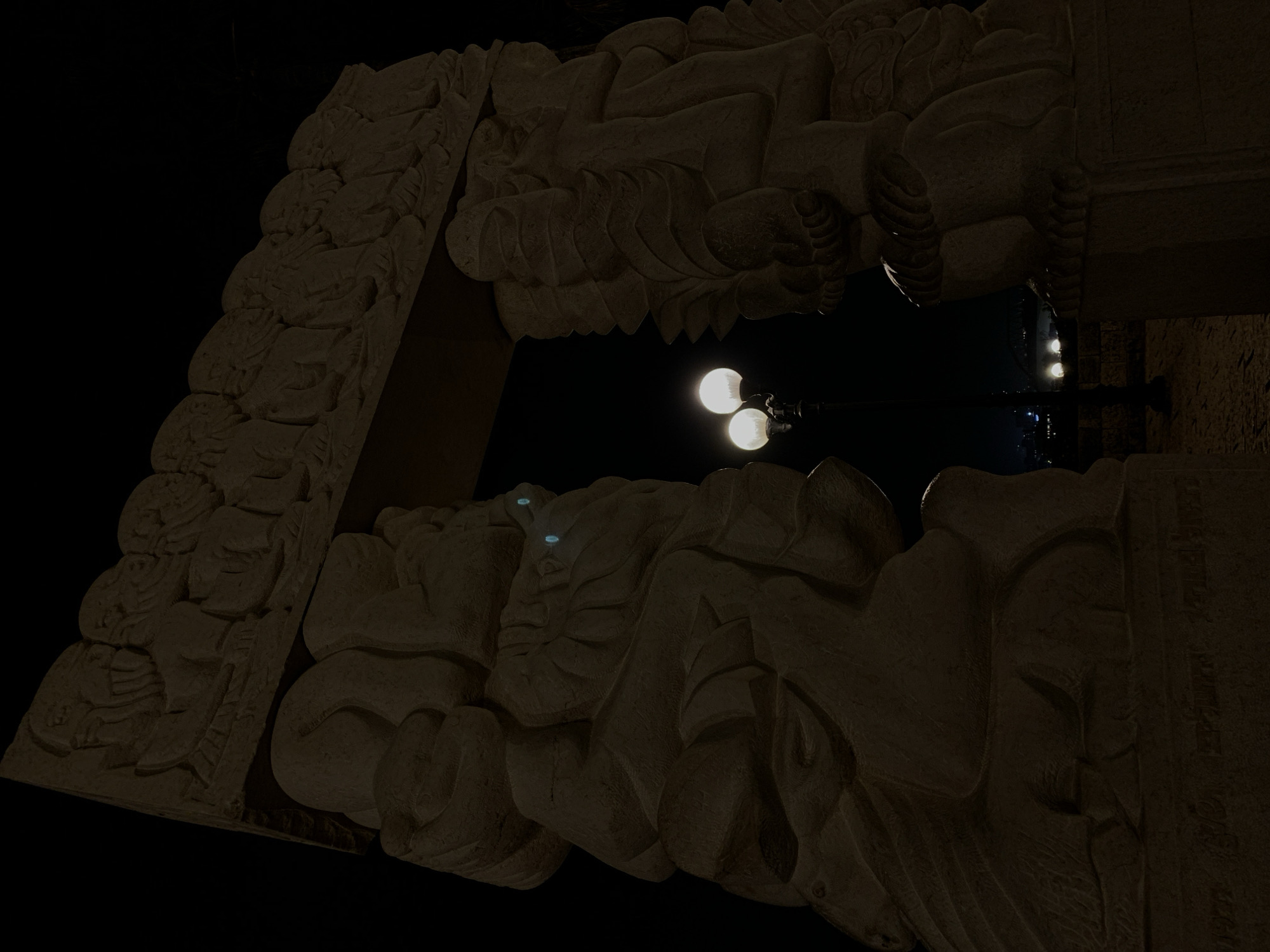 The image depicts a stone archway adorned with carvings, likely part of a building or monument. The archway frames a view of a street lamp with three illuminated bulbs against a dark sky. The carvings appear to be figurative, showing human or mythical forms. The location can be inferred to be in the city of Tel-Aviv, Israel.
