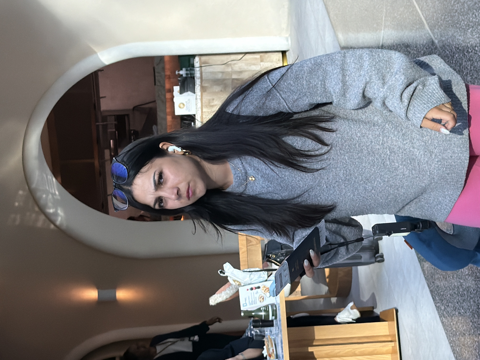 A young woman with long, dark hair and a grey sweater stands looking towards the camera with a confused expression. She is wearing blue-tinted sunglasses on her head and white earbuds. She holds a passport in her right hand and a small black electronic device attached to a lanyard in her left. She is wearing pink pants. In the background, a table with food and drinks is visible, along with other people and an arched doorway. The scene appears to be an indoor public space, possibly a cafe or airport lounge, with modern architecture and warm lighting. The presence of a passport suggests travel. The context of Doha, Qatar, indicates this is likely Hamad International Airport.