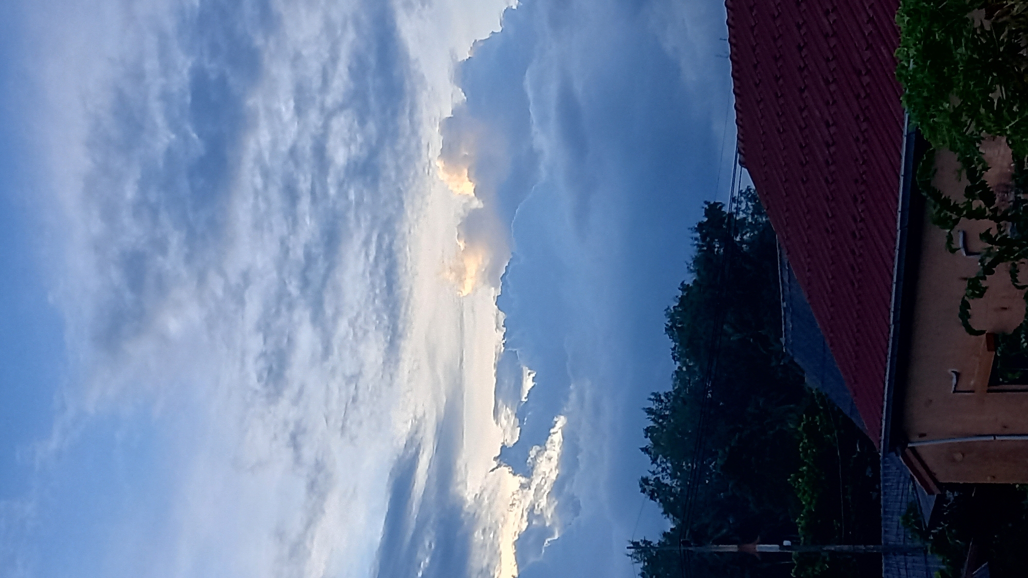 A vertical, outdoor, eye-level shot shows a cloudy sky over a residential area in Ko Pha-ngan District, Thailand. The sky is filled with large, dramatic clouds, some illuminated by sunlight peeking through, casting a golden glow. Below the clouds, a line of dark green trees is visible. In the foreground, the roofs of two houses are prominent: one with a red, corrugated tile roof and another with a dark, possibly slate, roof. A portion of a light-colored house is also visible, with a vine growing on its exterior. The scene appears to be captured during either sunrise or sunset, given the lighting conditions and the dramatic cloud formations. There are no people or animals visible in the image. The environment is residential, with evidence of typical housing structures.