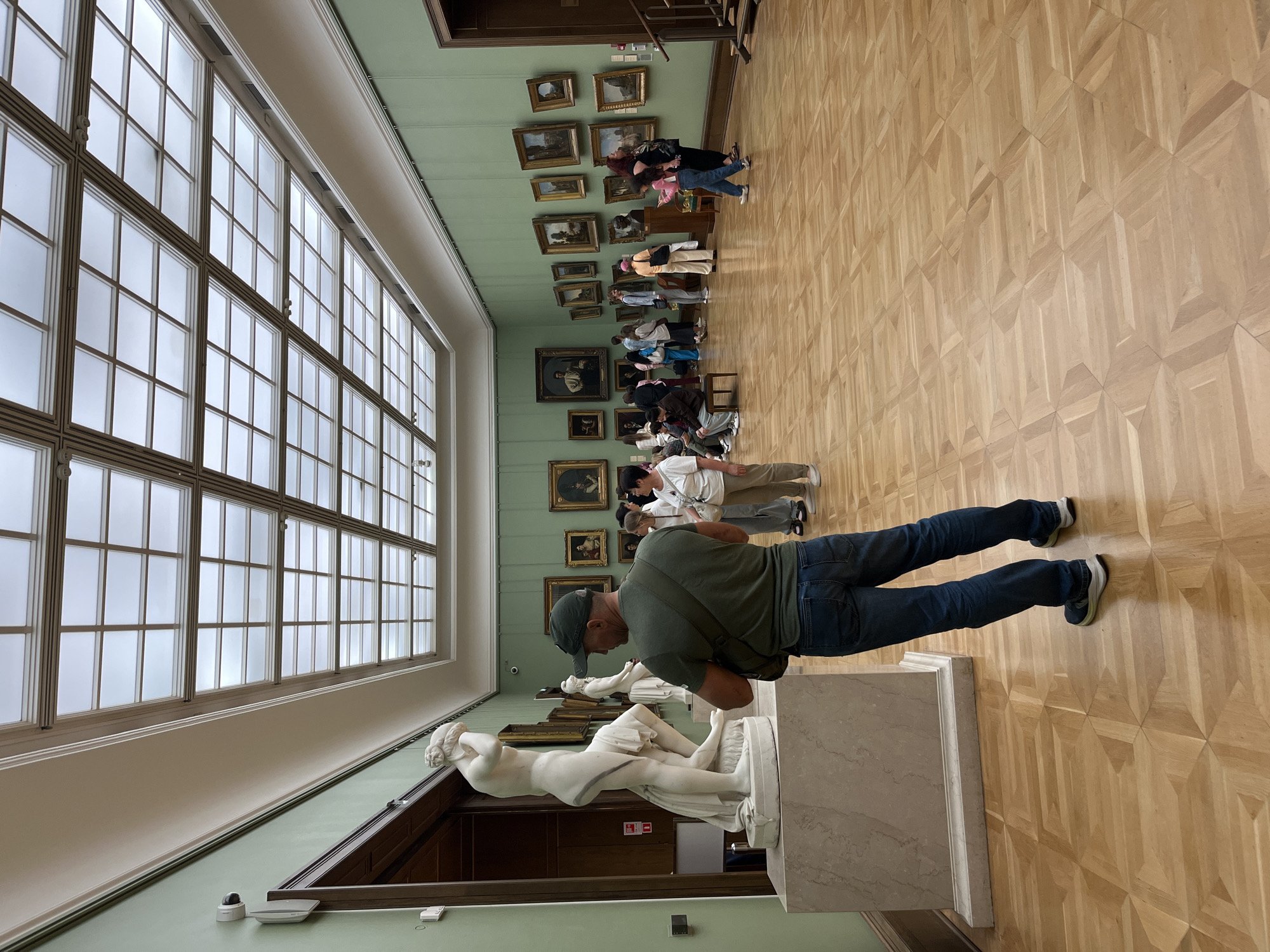 This image depicts a spacious art gallery with a high, grid-like ceiling made of glass panels. The walls are painted a light green, and they are adorned with numerous framed paintings and sculptures. A large marble statue of a female figure stands prominently on a pedestal in the foreground, to the left. In the middle ground, several people, including a man in a green shirt and cap facing away from the camera, are observing the art. Some are standing, while others are seated on benches. Further in the background, more individuals are scattered throughout the gallery, looking at various exhibits. The floor is made of polished wooden parquet with an intricate geometric pattern. The overall impression is that of a well-lit and busy museum environment. This scene is consistent with the interior of the Tretyakov Gallery in Moscow, Russia, known for its extensive collection of Russian art.