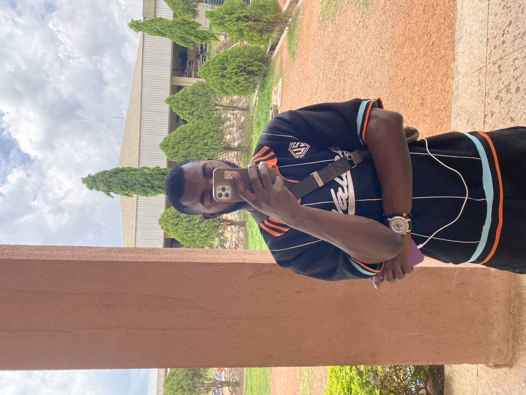 A man is outdoors, likely in Gwong, Nigeria, taking a selfie with his phone. He's wearing a black shirt with orange and blue accents, a watch on his left wrist, and a shoulder bag. He is holding a purple object in his left hand. The background shows a building, greenery including trees, and a cloudy sky. He appears to be standing next to a beige concrete pillar.