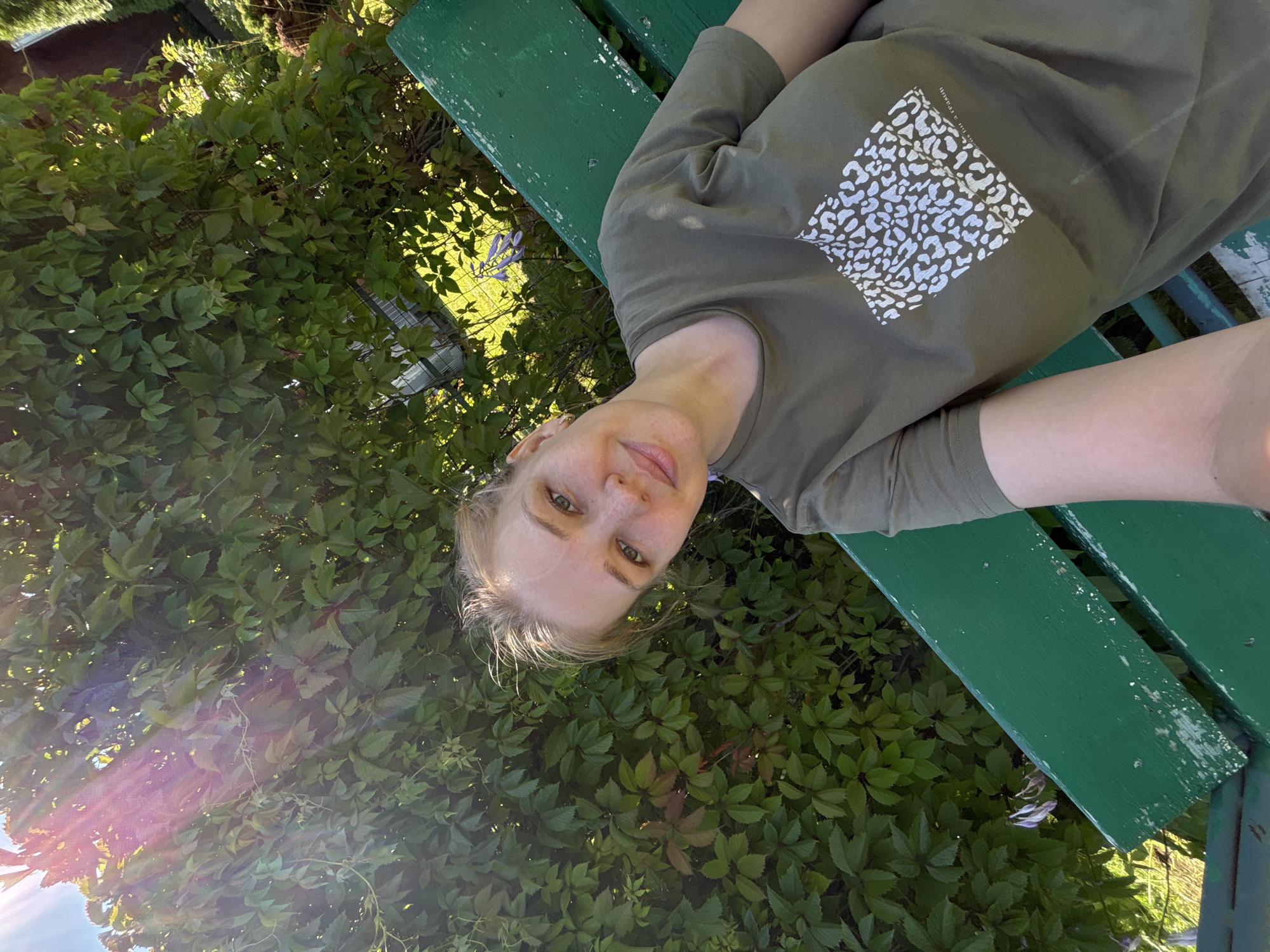 A young woman with blonde hair and freckles is sitting on a green wooden bench in front of a dense green hedge. She is wearing a green t-shirt with a white patterned square on it. The sun is shining brightly, casting lens flares on the left side of the image. The background shows more greenery and what appears to be a garden with some faint purple flowers. The setting is outdoors, likely a garden or park, and the presence of a wooden bench suggests a public or private outdoor seating area. The location is identified as Dunilovo, Russia.