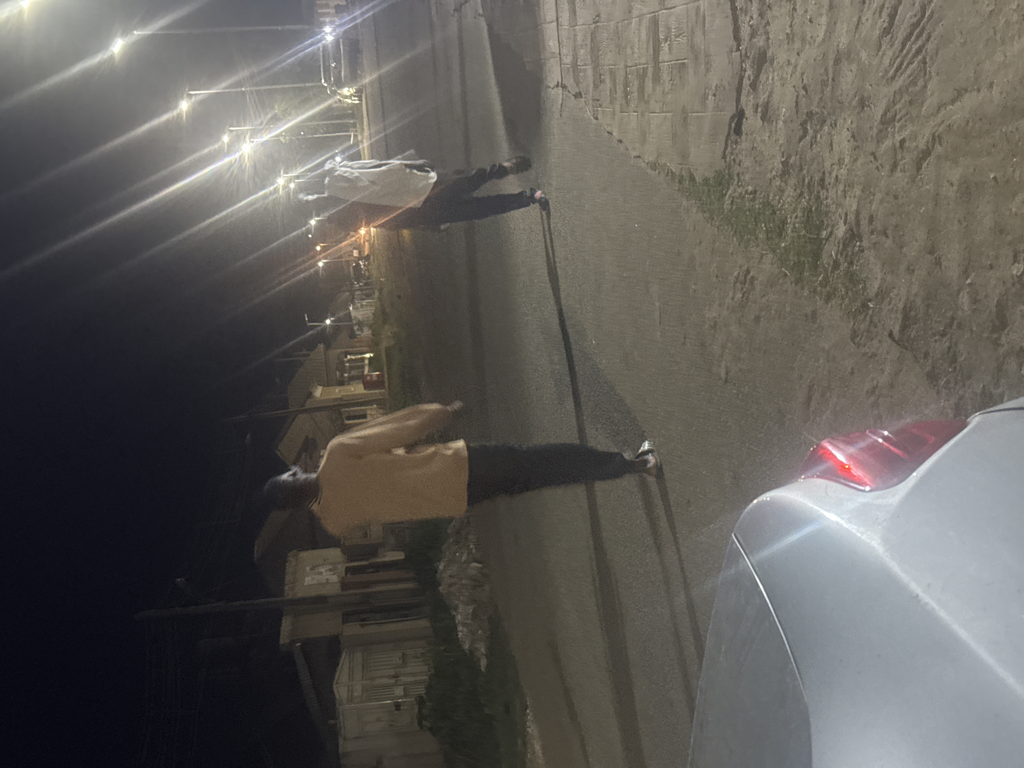 The image shows a nighttime street scene in Abuja, Nigeria. Two individuals are walking on a paved road under streetlights. The person in the foreground is wearing a light-colored long-sleeved shirt and dark pants, walking away from the camera. The second person, further down the road, is wearing a white garment and dark pants, also walking away from the camera. The environment includes residential buildings on the left side of the road and parked vehicles. The road surface appears to be asphalt. The lighting from the streetlights creates strong lens flares and highlights. A silver car is partially visible in the foreground on the left. The ground on the right side of the road appears to be a mix of dirt and pavement.