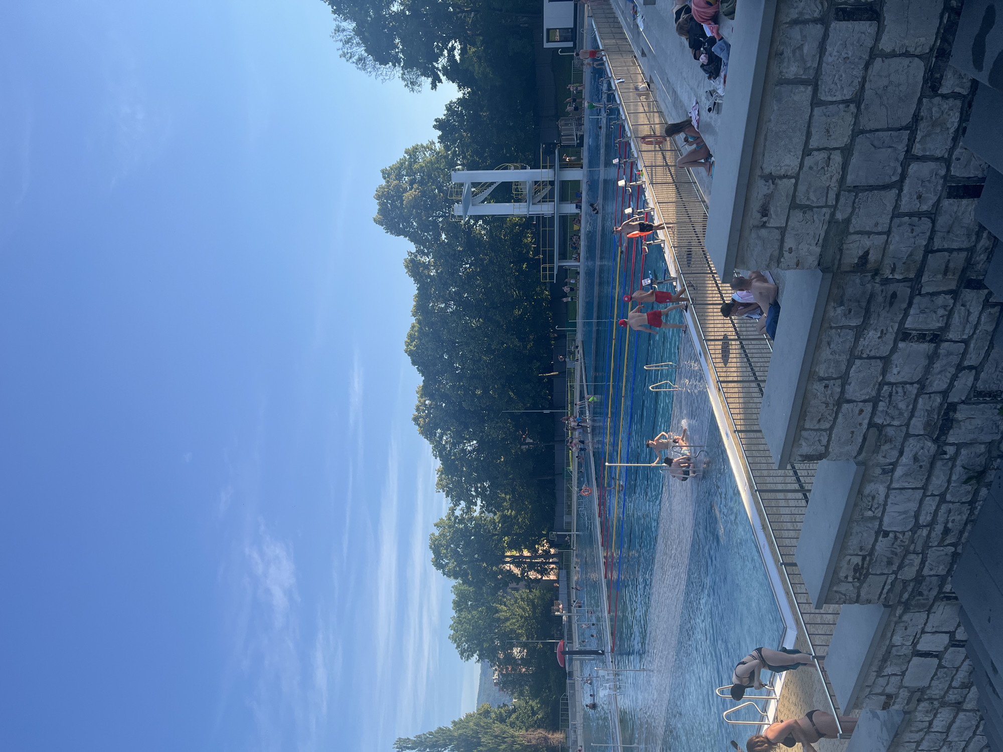 The image depicts an outdoor swimming pool complex on a sunny day. The pool is large and rectangular, divided into lanes by colorful ropes. Several people are swimming and wading in the water. Some are sunbathing on the tiered stone seating area that overlooks the pool. In the background, there are lush green trees and a clear blue sky with a few wispy clouds. A diving tower stands prominently on one side of the pool. The overall atmosphere appears relaxed and recreational, typical of a summer day at a public pool.