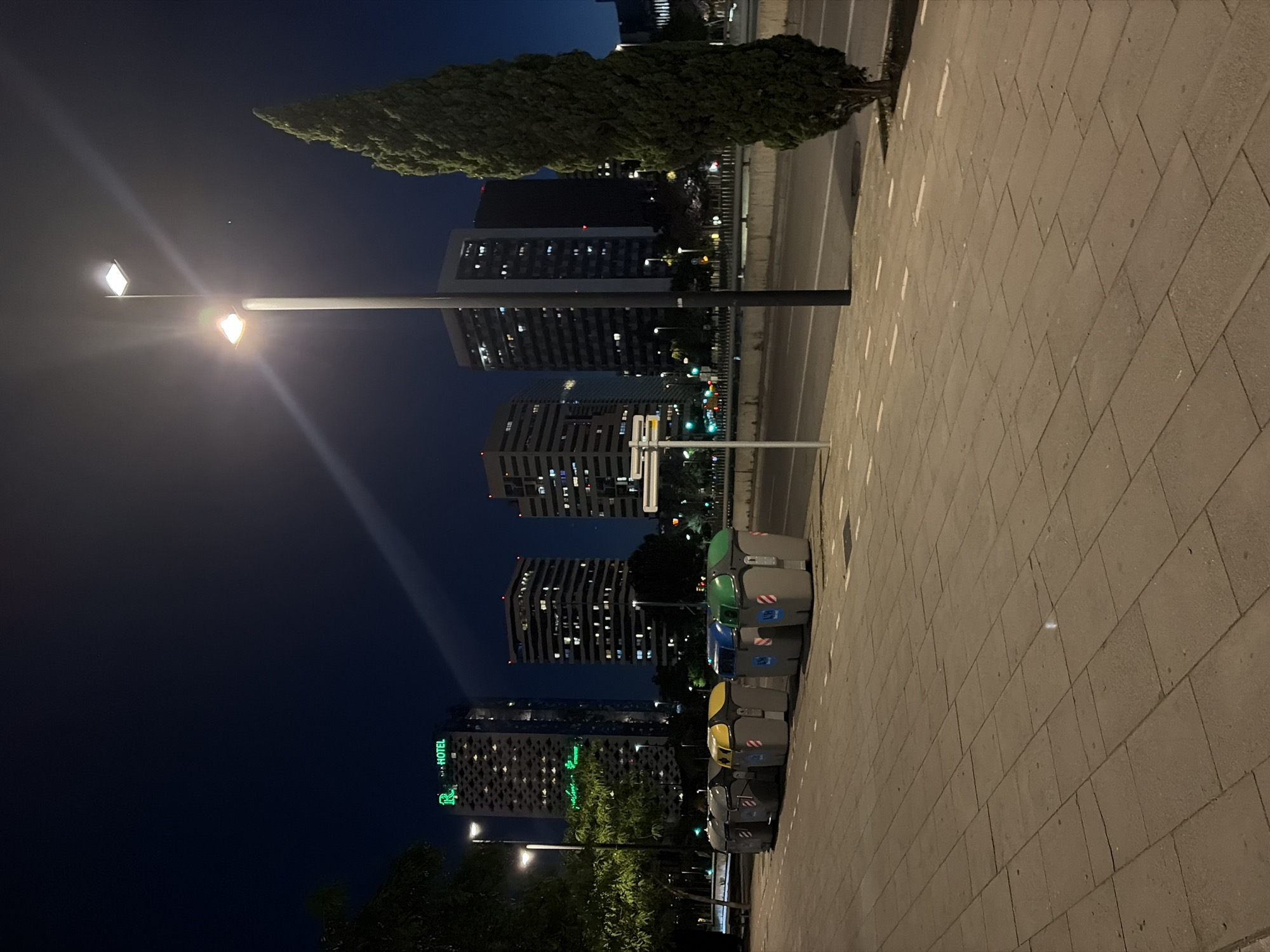 The image shows a nighttime street scene in L'Hospitalet de Llobregat, Spain. The foreground features a sidewalk paved with large, rectangular stones. Aligned along the sidewalk are several recycling bins of various colors (green, blue, yellow, grey). Behind the bins is a road and a line of trees. Beyond the trees, several tall buildings are visible, illuminated with lights. One building has a sign that says "HOTEL". A tall street lamp is positioned on the left side of the image, casting a bright light that obscures some of the sky. The sky is a dark blue/black color.