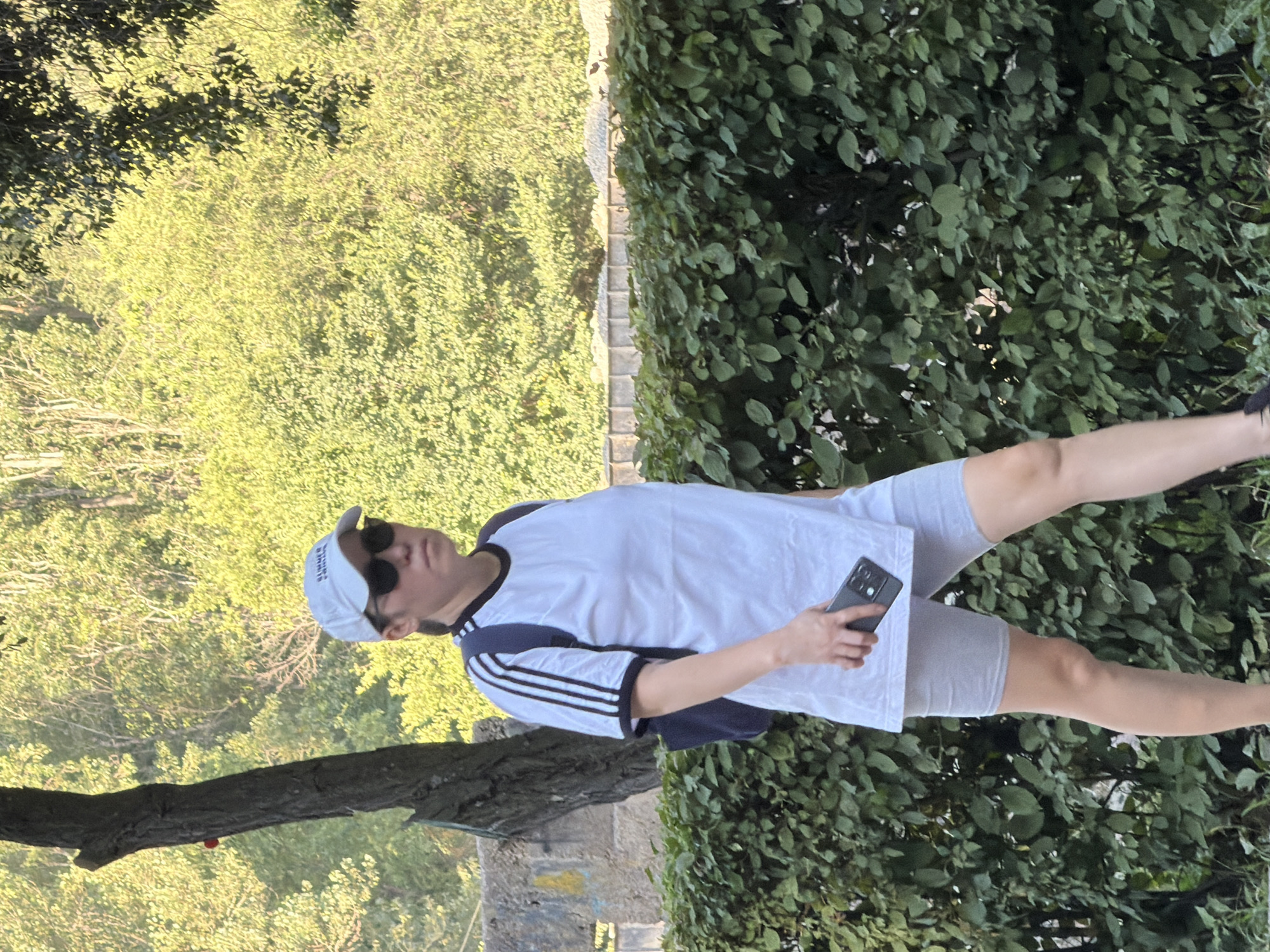 An individual is captured in a medium shot, walking outdoors and holding a black smartphone in their right hand. The person is wearing a white baseball cap, sunglasses, a white t-shirt with black stripes on the sleeves, and light gray biker shorts. They have a dark backpack on their left shoulder. The background consists of lush green foliage, including trees and a hedge, with a hint of concrete structures and dirt in the distance. The lighting suggests it is daytime, possibly late afternoon, with sunlight filtering through the leaves. The location is an outdoor, possibly park-like, setting.