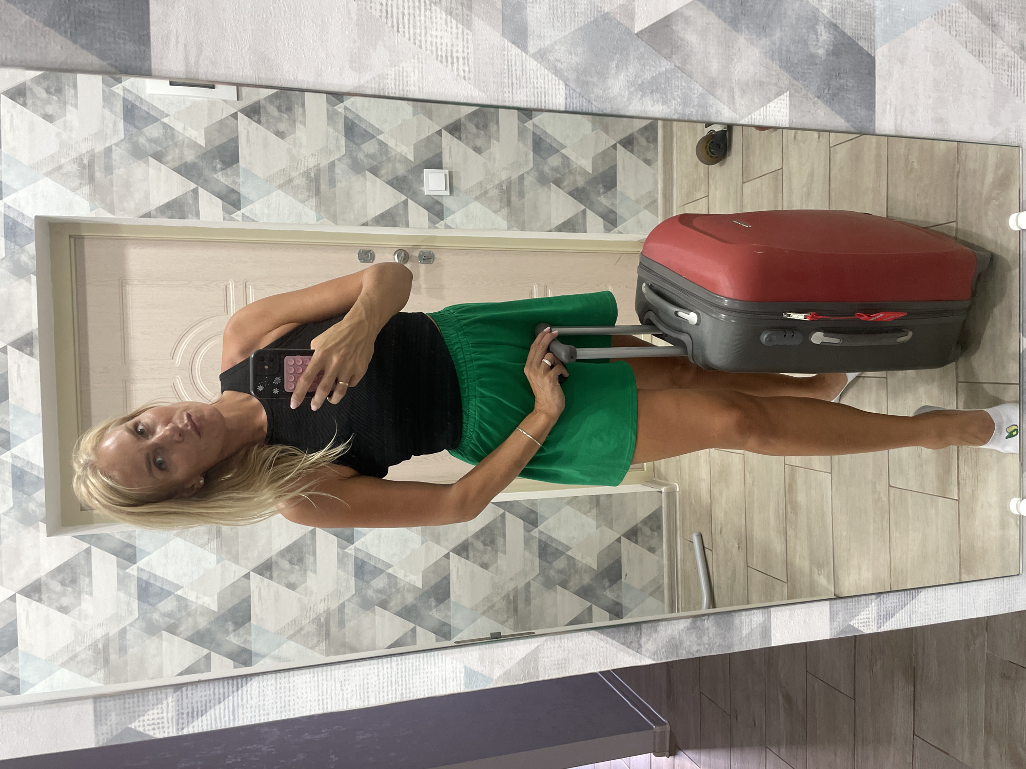 A woman with blonde hair and fair skin stands in front of a mirror, holding a smartphone in her right hand and a suitcase with her left. She is wearing a black tank top and a green skirt. The background shows a door and a wall with a geometric pattern. The floor is tiled. The scene appears to be indoors, possibly in a hallway or dressing area, in Novorossiysk, Russia.