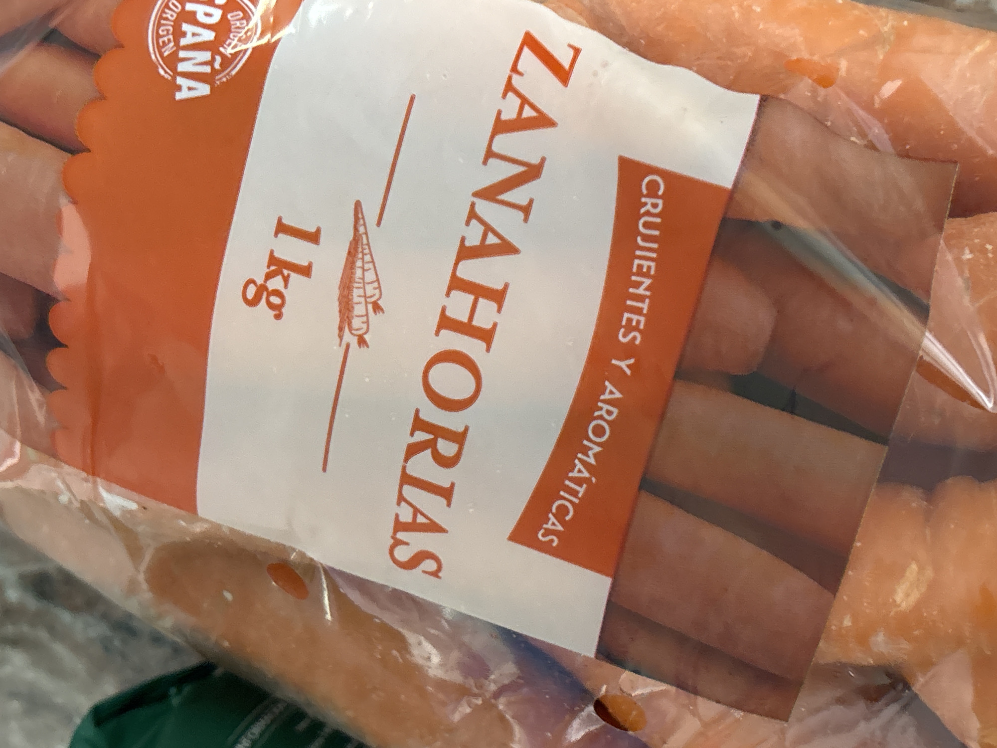 A plastic bag filled with carrots is shown. A label on the bag reads "ZANAHORIAS" in large orange letters, with "CRUJIENTES Y AROMÁTICAS" below it. To the top right of the "ZANAHORIAS" text, there is a circular stamp that says "ORIGEN" and "SPANA". The image does not provide any information that allows for the location in the city of Orihuela, Spain to be determined.