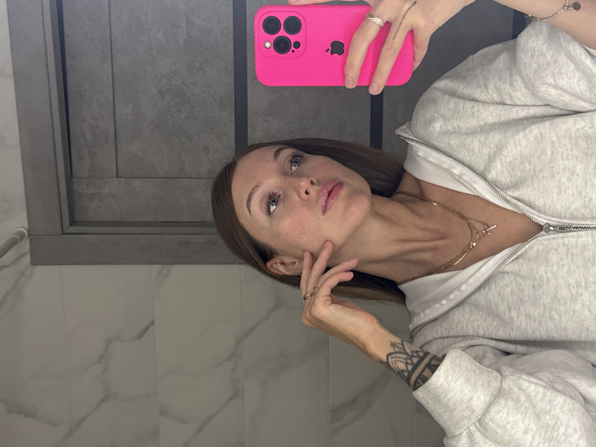 A woman is taking a selfie in a bathroom with gray tiles, while holding a pink iPhone. The location is likely a residence, possibly located in or near the city of Chaikovsky, Russia. She is wearing a light gray zip-up hoodie and a gold necklace. She has a tattoo on her left forearm. Her right hand is touching her face near her cheek.