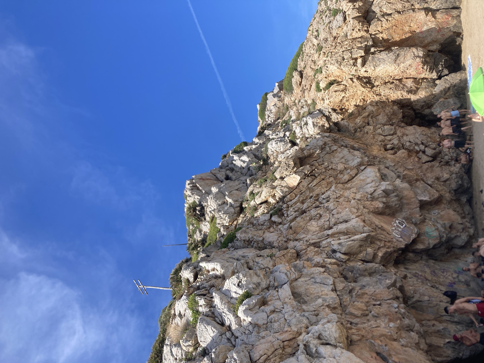 A high-angle, medium shot captures a rocky cliff face on a beach in Port De La Ginesta, Spain. The cliff, composed of layered, rough-textured rock in shades of tan, orange, and gray, dominates the left and center of the frame. Sparse green vegetation grows from crevices in the rock. At the top of the cliff, a wooden utility pole with two crossbars stands against a bright blue sky filled with wispy white clouds and a faint contrail. The lower right portion of the image reveals a sandy beach with a few people gathered near the base of the cliff, some of whom are partially obscured by the shadow of an overhang. Several individuals are standing and conversing, while others appear to be sitting or relaxing. A bright green umbrella is partially visible in the foreground, suggesting a recreational setting. Graffiti in white, red, and green is present on the lower sections of the cliff face.