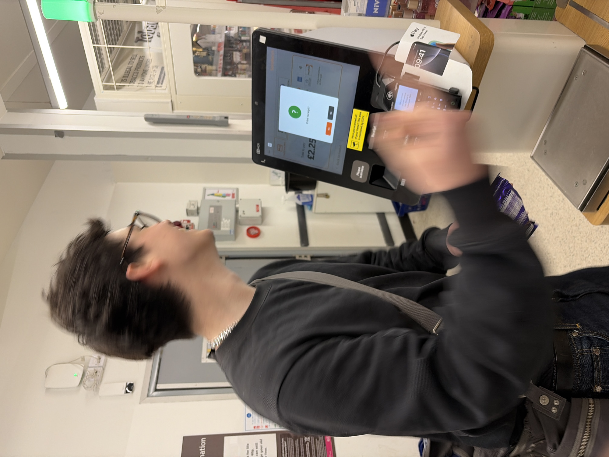 A young male with dark hair and glasses is at a self-service checkout in a supermarket, likely in London, United Kingdom, as inferred from the signage in the background. He is wearing a black sweater and jeans. His arm is raised, likely interacting with the checkout screen, which displays a prompt asking if he wants to print a receipt. The total to pay is displayed as £2.25. The checkout station includes a payment terminal and a scale. Shelves with various products are visible in the background. The environment appears to be brightly lit. Signage nearby cautions that it is illegal to sell tobacco products to anyone under 18.