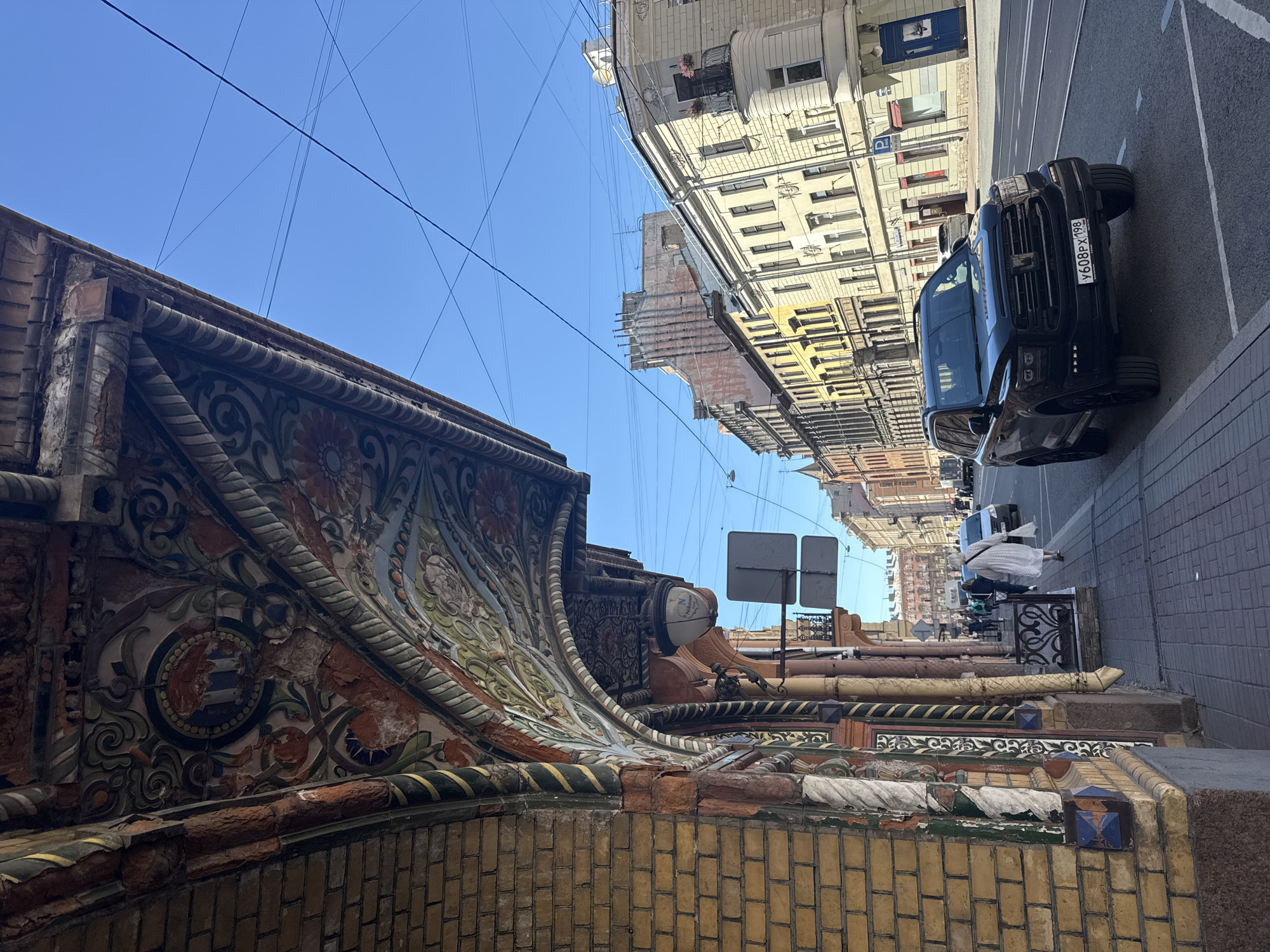The image shows a street scene in Saint Petersburg, Russia, with a focus on the ornate architecture of a building. The photograph is taken from a low angle, looking up at a decorated facade and then extending to the street and buildings across.
The foreground features a close-up of the building's exterior, characterized by intricate tilework and brickwork. The decorative elements include floral patterns, geometric designs, and textured surfaces in shades of orange, green, white, and blue. Some of the tiling appears weathered and damaged, suggesting age.
Beyond the ornate facade, the scene opens up to a street with a black SUV parked on the right side. The car's license plate is visible and reads "Y608PX 198". The street is paved, and tram tracks are visible running down the middle. Across the street, several multi-story buildings line the road, displaying a variety of architectural styles common in Saint Petersburg, with numerous windows and balconies.
The sky above is a clear, bright blue, indicating a sunny day. Several power lines crisscross the sky, a common sight in urban environments. There are also two square, gray signs mounted on poles on the right side of the street.
While no specific event is clearly depicted, the presence of the parked car and the general street activity suggest a normal day in the city. The overall mood is one of urban exploration, highlighting the rich architectural heritage of Saint Petersburg. The time of day appears to be daytime, likely late morning or early afternoon, given the bright sunlight.