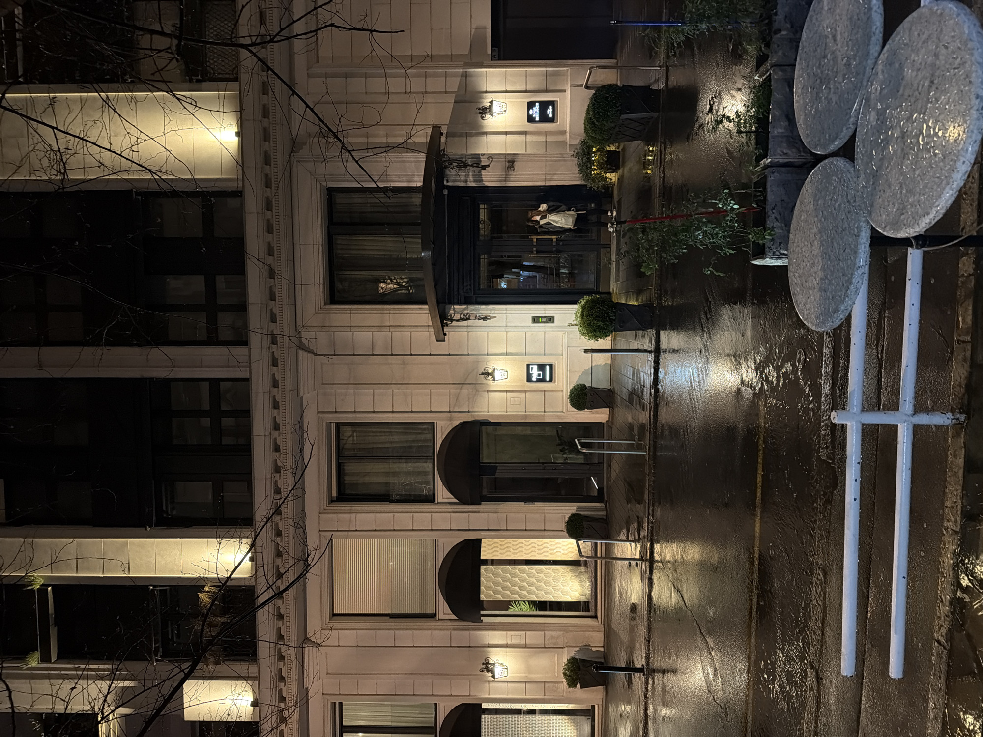 An exterior forensic analysis of the image reveals a nighttime scene in front of the Regent Hotel, located at Kralja Milana 34, Belgrade, Serbia. The ground, including the street and sidewalk, is visibly wet, indicating recent precipitation, with numerous reflections of artificial light sources.
The central subject is a multi-story building with a light-colored, possibly stone or stucco, facade exhibiting classical architectural elements such as pilasters, cornices, and decorative friezes. The building is illuminated by warm-toned exterior lighting, including wall-mounted sconces flanking the entrances and uplights on the upper facade.
The main entrance, positioned slightly to the right of the image center, features a dark, ornate canopy over dark double doors. The number "34" is affixed to the wall next to the doors. To the right of the main entrance, a square sign with "REGENT" is mounted on the facade, below another ornate wall sconce. To the left of the main entrance, two additional arched doorways, each with a dark awning, are visible. One of these doorways reveals an interior with a distinct patterned wall covering.
One individual is observed standing directly in front of the main entrance doors. They are wearing a light-colored top (possibly a hoodie or jacket), dark pants, and athletic shoes, carrying a light-colored backpack. The individual appears to be looking down at an object held in their hands, potentially a mobile device.
Several potted evergreen shrubs are placed along the building's facade near the entrances. Bare tree branches extend across the upper portion of the image, silhouetted against the building. In the foreground, three wet, round, light-colored outdoor tables are partially visible, positioned behind a white, two-bar railing. Dark street bollards are sparsely placed along the curb line. The wet asphalt of the street shows a faint yellow line.