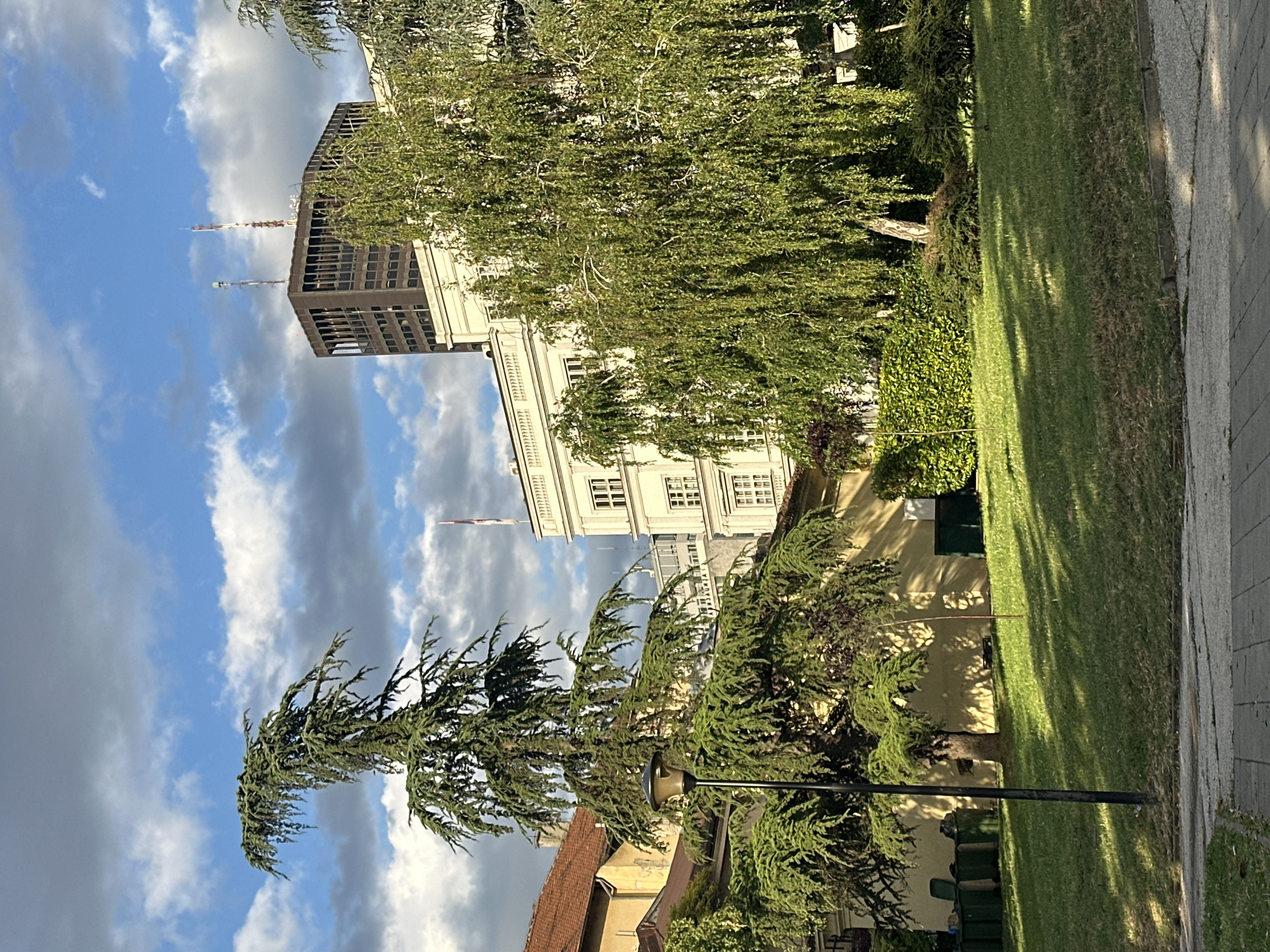 In the heart of Belgrade, Serbia, a serene park setting unfolds beneath a dramatic sky. The scene is dominated by lush greenery, with a mature deodar cedar tree in the foreground, its branches gracefully arching over a well-maintained lawn. To its right, a weeping birch tree adds a delicate, cascading element to the landscape. Behind the trees, a modern, cream-colored building with classical architectural details stands tall, its windows reflecting the sky. Further in the background, a dark, imposing skyscraper with vertical window elements pierces the clouds, topped with antennas. The park features a paved walkway in the foreground and a grassy slope leading up to the buildings. Several green waste bins are visible near a building on the left. The time of day appears to be late afternoon or early evening, as evidenced by the dappled sunlight and long shadows cast across the grass. The overall impression is one of urban tranquility, where nature and architecture coexist harmoniously.