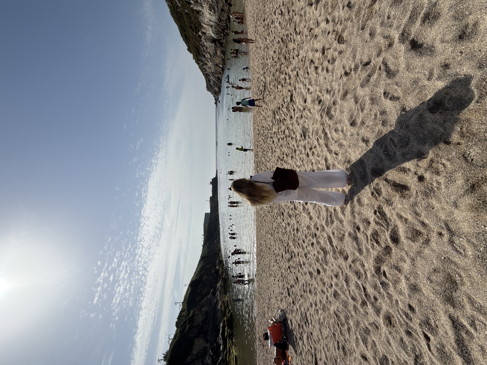 The image shows a sunlit beach scene, likely in A Coruña, Spain, with a body of water and hilly terrain in the background. The foreground is dominated by sand, with footprints and a long shadow cast by a woman standing with her back to the camera. She is wearing white wide-leg pants and a white top, carrying a dark bag over her shoulder.
In the middle ground, the water is populated by many people swimming and wading. Some are standing further out, while others are closer to the shore. Children are visible among the adults. On the left side of the sand, two people are seated in red chairs, one lying down and the other reclined.
The background features a calm body of water that appears to be a bay or cove, framed by rocky, vegetated hills on either side. A man-made structure, possibly a building or fortification, is visible on the far right hill. The sky is bright blue with scattered clouds and the sun is a strong, bright presence in the upper left corner. The overall impression is a busy, sunny day at a popular beach.