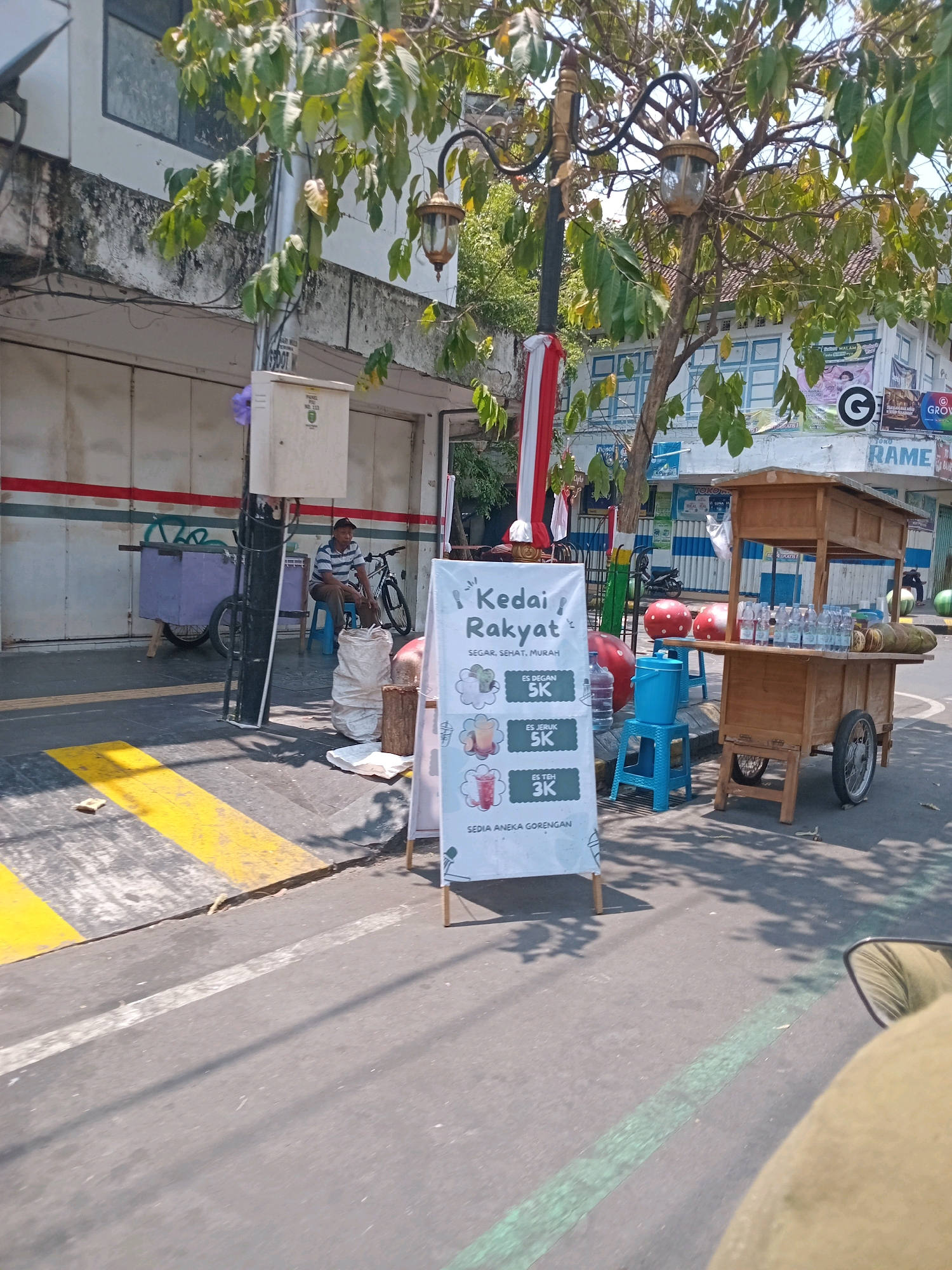 A medium shot captures an outdoor scene on a sunny day, likely in a city. In the foreground, a white A-frame sign displays "Kedai Rakyat" (People's Stall) with the tagline "SEGAR, SEHAT, MURAH" (Fresh, Healthy, Cheap) and prices for drinks like "ES DEGAN 5K," "ES JERUK 5K," and "ES TEH 3K" (Coconut ice, Orange ice, Tea ice). Below these are the words "SEDIA ANEKA GORENGAN" (Assorted Fried Snacks available). To the right of the sign, a wooden pushcart vendor stall is set up, displaying bottled drinks and cut coconuts on its top shelf. A blue plastic bucket and two red spheres sit on blue stools near the stall. In the background, a man in a striped shirt sits on a blue stool next to a bicycle and a purple wheeled cart. Buildings with closed doors line the street, and trees with green leaves cast shadows. A lamppost with a decorative lantern stands near the sign. The street has yellow and white markings, indicating a pedestrian crossing.