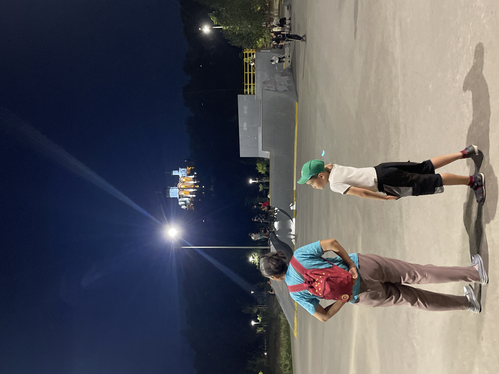 In the city of Almaty, Kazakhstan, at night, an outdoor skatepark is illuminated by artificial lights. In the foreground, two figures stand facing away from the camera, looking towards the skatepark. The figure on the left, an adult, wears a blue t-shirt, beige pants, and a red backpack with yellow star patterns. The figure on the right, a child, wears a white t-shirt, black shorts with grey panels, and a green baseball cap.
In the mid-ground, several individuals are engaged in skateboarding and scootering on ramps and flat surfaces. The skatepark features various concrete ramps and structures, with yellow railings visible in the background. Beyond the skatepark, a dense line of trees forms a dark silhouette against the night sky.
In the far distance, atop a hill, a brightly lit building, appearing to be a church or a monastery with multiple domes, stands out against the dark sky. The building is illuminated with white and yellow lights, and a bright beam of light emanates from a streetlamp in the skatepark, projecting upwards towards the sky and past the illuminated structure. The sky is a deep, dark blue, indicating nighttime. The ground of the skatepark is a smooth, light-colored concrete surface.