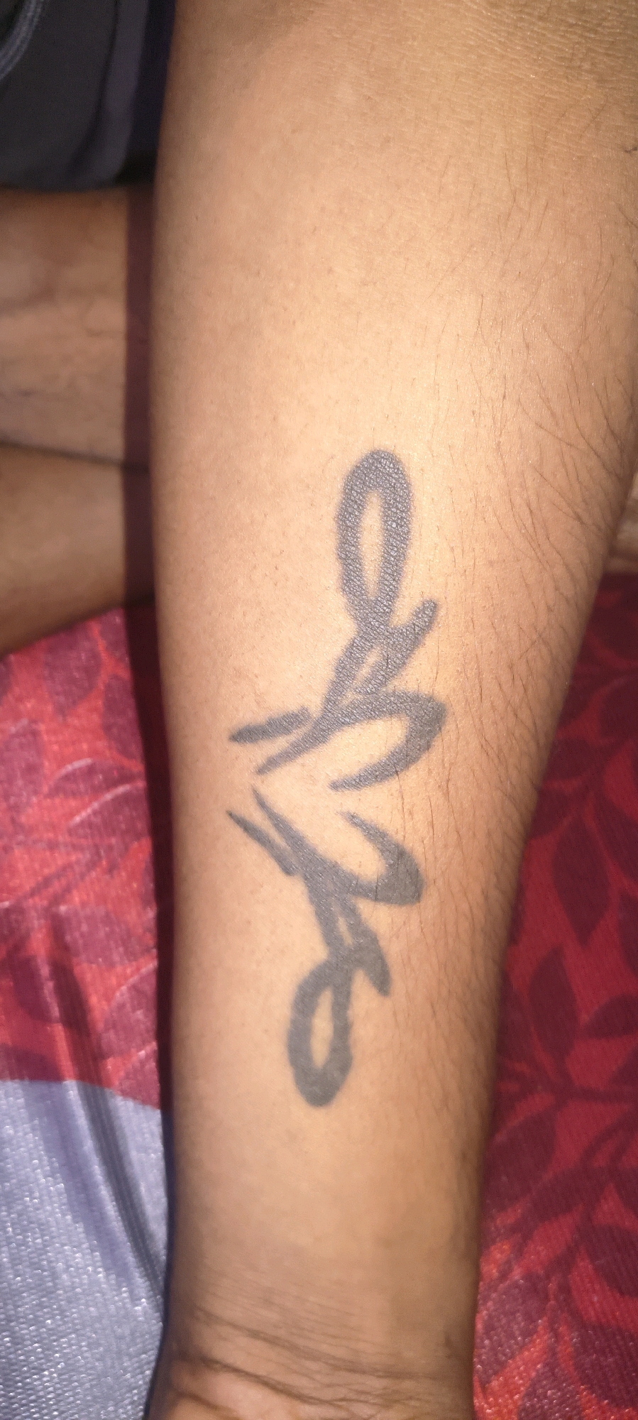 The image shows a close-up view of a tattoo on a person's forearm. The tattoo appears to be in black ink and is stylized, possibly depicting letters or abstract shapes. The skin has fine hairs visible. The background consists of textured fabric, predominantly in shades of red and purple, with some blue fabric on the left side. The setting appears to be indoors, possibly a home environment. No specific location in Bengaluru, India is identifiable from the image itself.