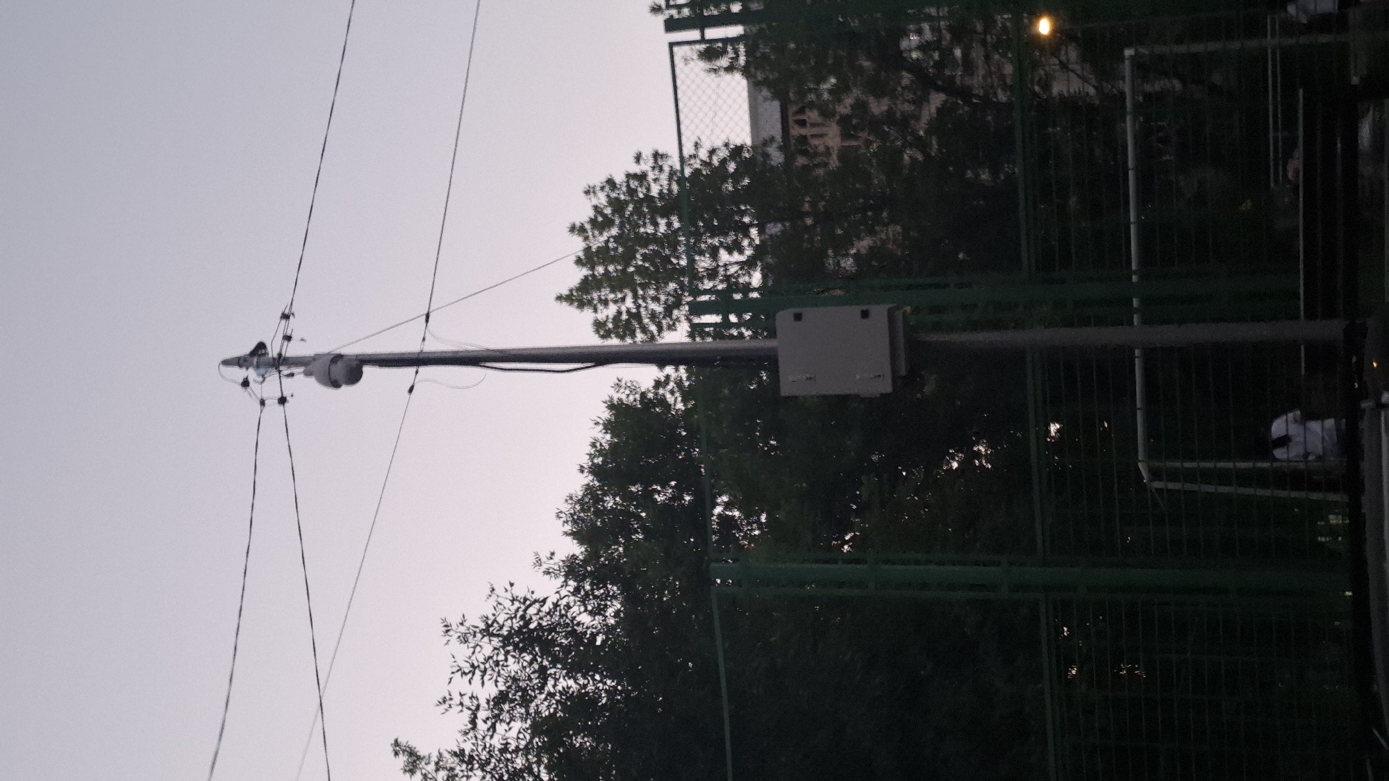 The image shows an outdoor scene with a utility pole as the central focus. Attached to the pole are electrical wires and a security camera. Below the camera, a gray electrical box is mounted on the pole. The background consists of lush green trees, a green metal fence, and what appears to be a sports field with a goalpost. The sky is overcast, suggesting it may be evening or a cloudy day. In the lower portion of the image, a person wearing a white shirt is visible behind the fence. The scene suggests a public area within the city of Shymkent, Kazakhstan, possibly a park or recreational facility.