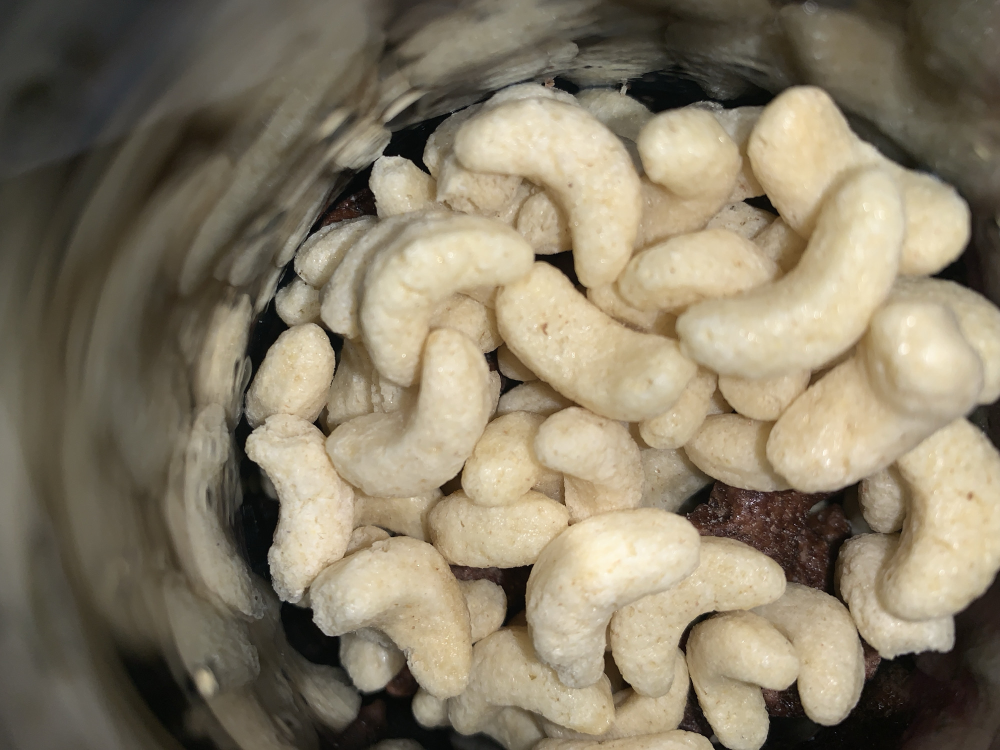 A close-up, indoor shot focuses on the inside of a cereal container, partially filled. The main subject is a heap of crescent-shaped, light-colored cereal pieces. Underneath, there are some darker, irregularly shaped pieces of chocolate cereal. The container's edge is blurry and out of focus on the left. The image details suggest that the container has been opened, and cereal has been consumed. There is no indication of location in Rafin Par, Nigeria, or people involved.