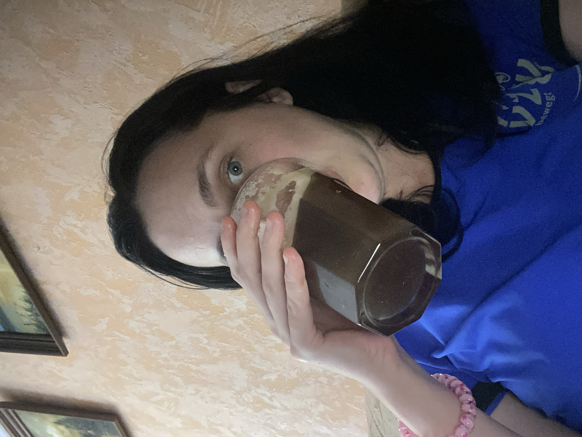 A close-up, waist-up, selfie-style, indoor shot depicts a woman with dark hair and light skin taking a drink from a faceted, clear glass filled with a dark liquid. She is wearing a bright blue t-shirt with a graphic design on the right side of her chest that appears to be text and symbols. On her left wrist, she is wearing a pink beaded bracelet. The background wall is textured and painted in a peachy, off-white color. A framed picture is visible on the wall to the upper left of the frame, depicting a landscape scene. The lighting is artificial and casts soft shadows. The woman's face is partially obscured by the glass and her hand. Her blue eyes are visible and looking towards the camera.