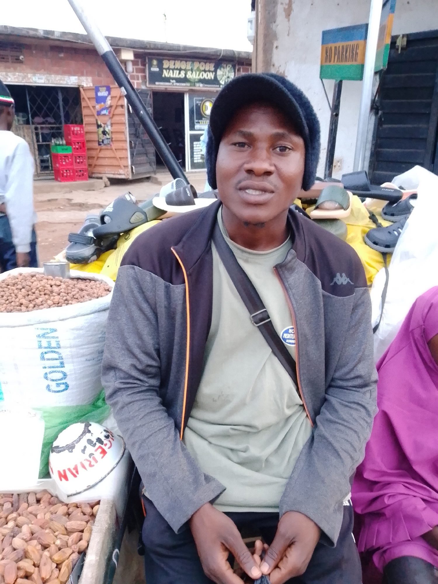 The image shows a man in a black beanie and a grey jacket with an orange zipper trim, sitting in what appears to be an outdoor market. In front of him are two large sacks, one with "GOLDEN" printed on it in blue letters, filled with small, round, brown objects, likely nuts or seeds. To the left of the sacks is a wooden tray filled with more of the same brown items. A white object with red lettering that reads "GERMAN" is also visible on the edge of the tray. Behind the man, a woman in a purple garment is visible. In the background, there is a building with signs that read "DENGE POSE NAILS SALOON" and "NO PARKING". There are also red and green crates stacked near the building. The overall setting suggests a street market environment in an urban area.