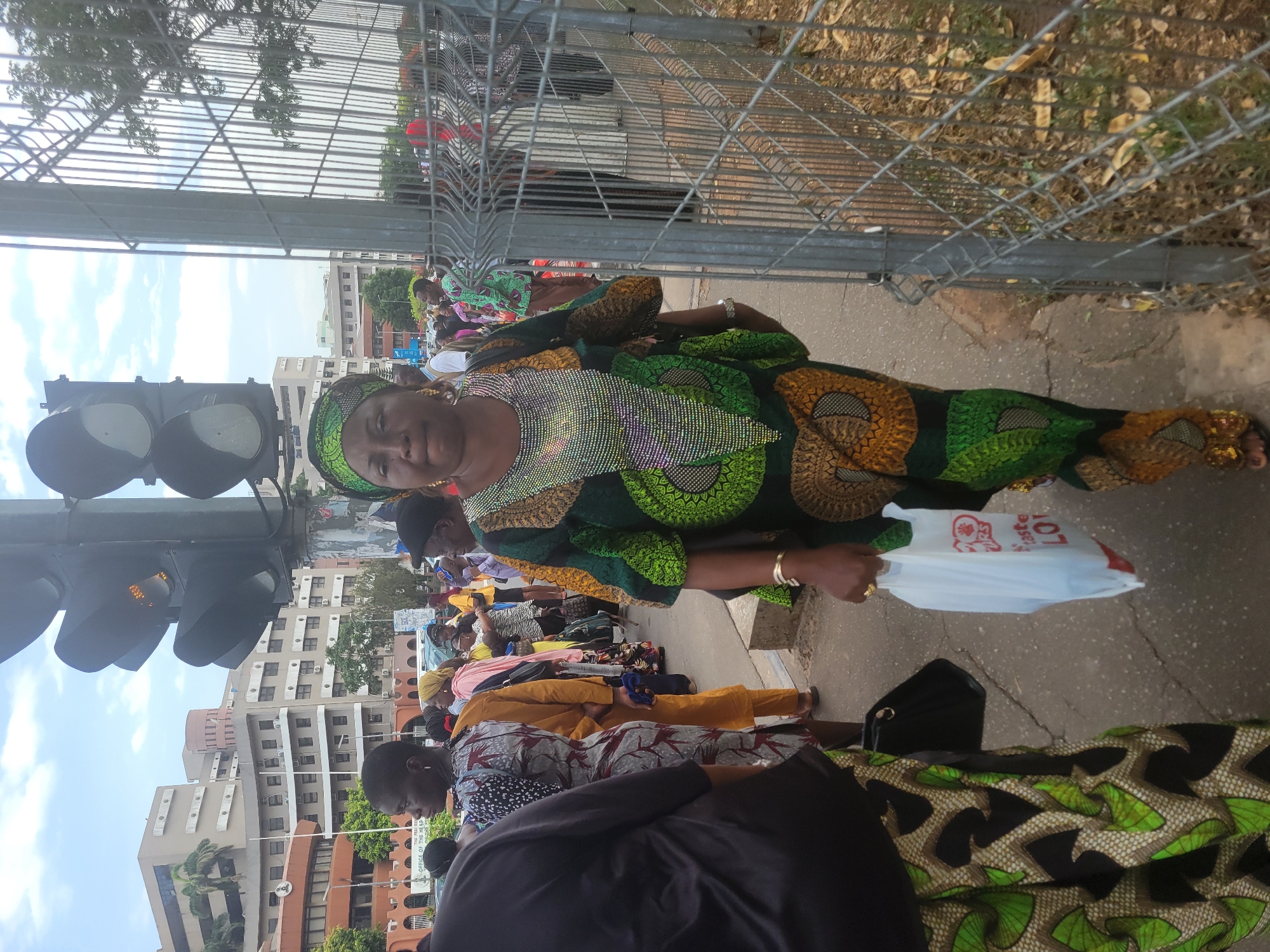A woman in traditional green and gold African attire stands on a sidewalk next to a fence and traffic lights in Abuja, Nigeria. She holds a white plastic bag. Other people, also dressed in African attire, are nearby. Buildings are visible in the background.