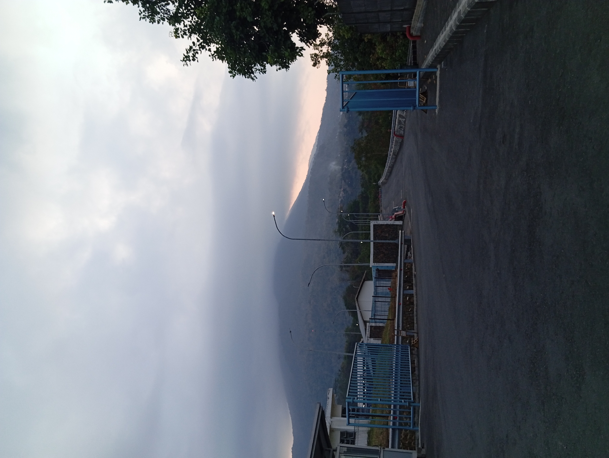 The image depicts an overcast day in Sumber Suko, Indonesia. In the foreground, an asphalt road curves towards the right, bordered by a concrete curb on the right and a metal guardrail with a stone embankment on the left. A blue metal gate is positioned on the left side of the road. Beyond the gate, a low-rise building with white walls and a dark roof is visible. Several streetlights line the road, with the nearest one on the left illuminated.
In the middle ground, the road continues to ascend, with more streetlights interspersed. A large, dark green tree is situated on the right side of the road, its branches extending towards the sky. Further in the background, a mountain shrouded in clouds dominates the landscape. The lower slopes of the mountain are covered in dense vegetation. A small, white building with a dark roof is nestled amongst the trees on the left side of the road.
The sky is filled with heavy, gray clouds, obscuring the sun and creating a muted lighting condition. The overall atmosphere is calm and serene. A single individual, wearing dark clothing, is seated on the ground near the guardrail on the left side of the road.