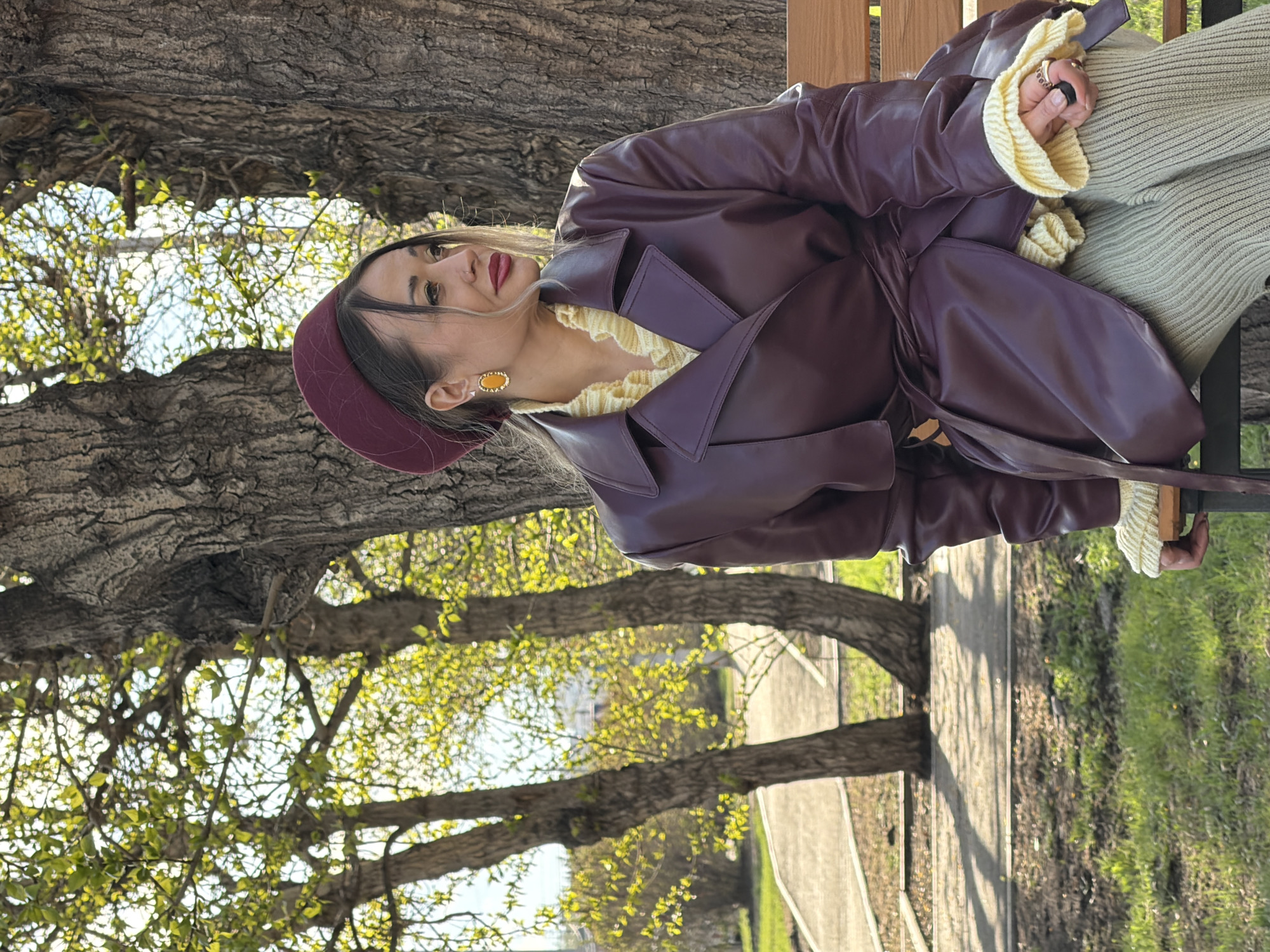 A woman is sitting on a park bench beneath trees, likely in Krasnoyarsk, Russia. She wears a burgundy beret and a matching leather coat with a belt. Her sweater is yellow with ruffled edges, and she wears a ring on her left hand. A paved path and green grass are visible in the background.