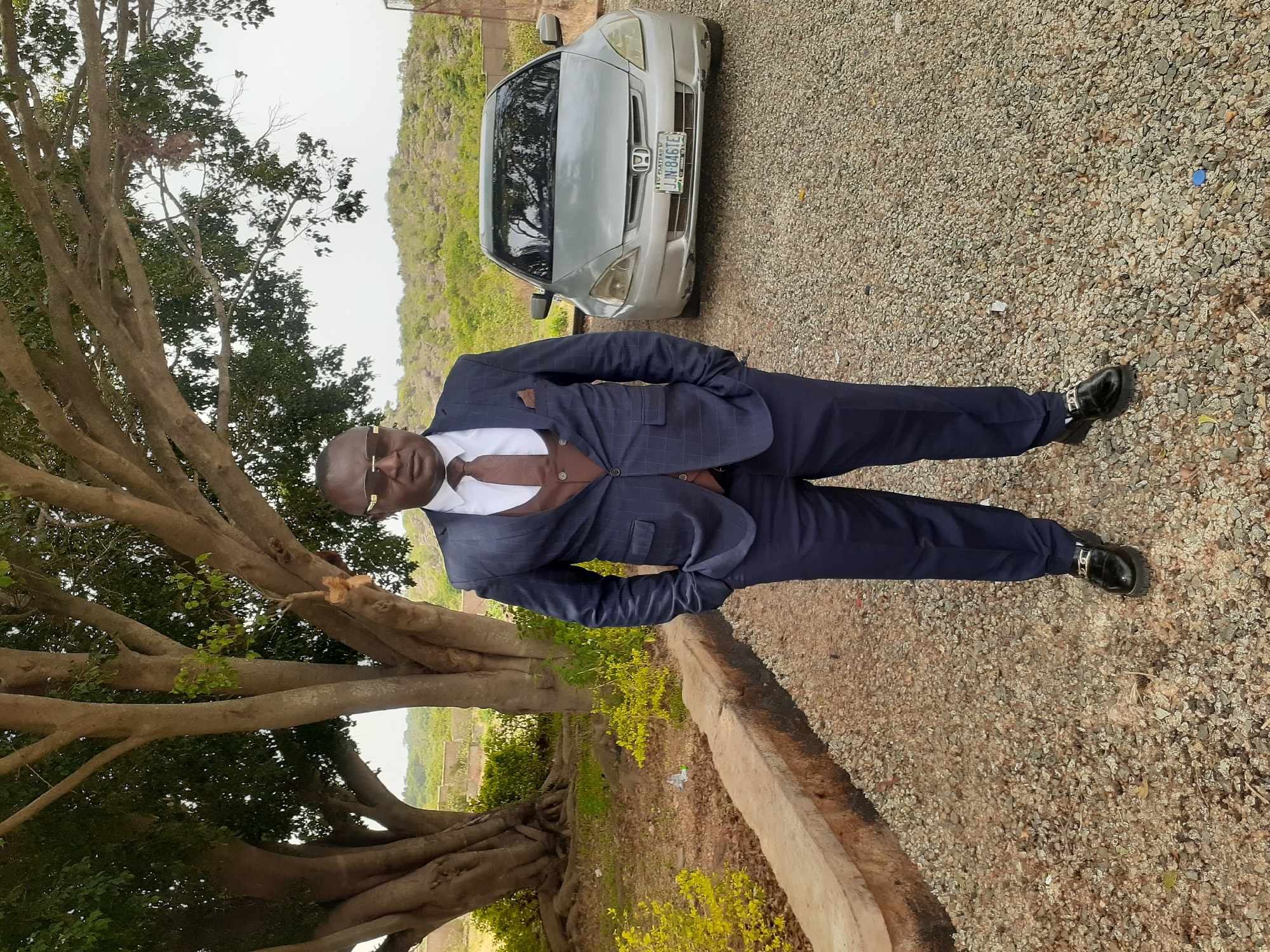 A full-length portrait depicts a man standing outdoors on a gravel surface, looking directly at the camera with a neutral to slight smile. He is formally dressed in a dark blue pinstripe suit jacket, matching trousers, a white shirt, a brown vest, and a brown tie. He completes his attire with dark shoes featuring metallic buckles and dark sunglasses. His hands are casually placed in his suit pockets.
To the left of the man, a large, mature tree with thick, gnarled branches and prominent exposed roots dominates the foreground. Lush green foliage is visible on the tree and scattered on the ground. Behind the man and slightly to his right, a silver sedan, identifiable as a Honda by its emblem, is parked on the gravel. The car's license plate, "JJM 846TE" and "PLATEAU," is clearly visible, indicating Plateau State, Nigeria, as a potential location. The car's front bumper shows some minor cosmetic damage.
In the background, rolling hills or mountains covered in sparse green vegetation rise under what appears to be an overcast or bright daytime sky. The ground throughout the scene is composed of small, light-colored gravel. The setting suggests a serene outdoor environment, possibly a rural or semi-rural area.
