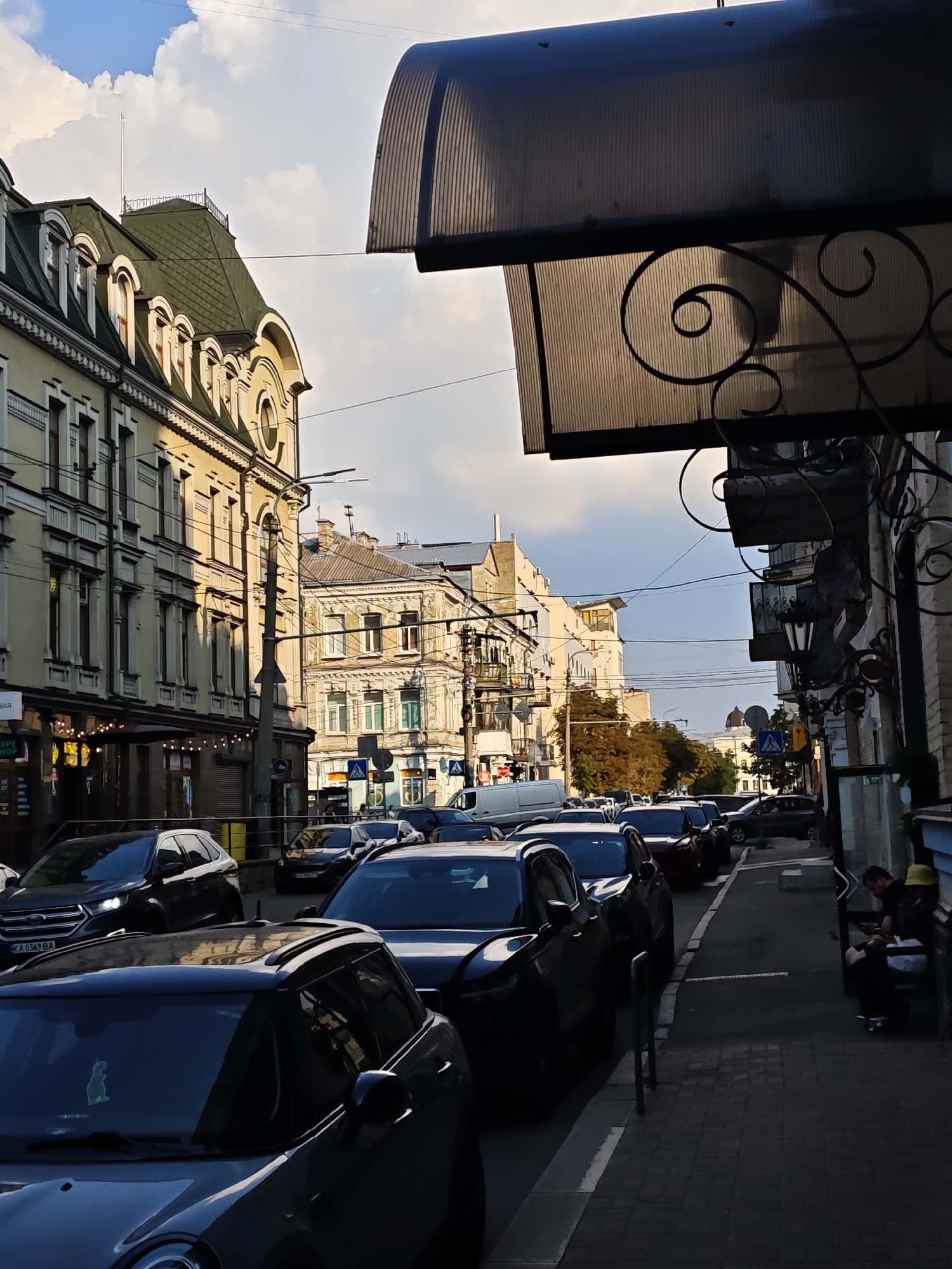 This image depicts a street scene in Kyiv, Ukraine, likely in a historic district given the architecture. On the left, a light-yellow building with ornate detailing and a green mansard roof with dormer windows dominates the foreground. A street lamp and power lines are visible in front of it. The street is lined with parked cars, mostly dark-colored sedans and SUVs, facing away from the viewer. On the right side of the street, a sidewalk features a person sitting on a low wall, looking at their phone, and another person next to them wearing a yellow hat. Above the sidewalk, a translucent awning with decorative ironwork extends from a building. The sky above is partly cloudy with patches of blue.