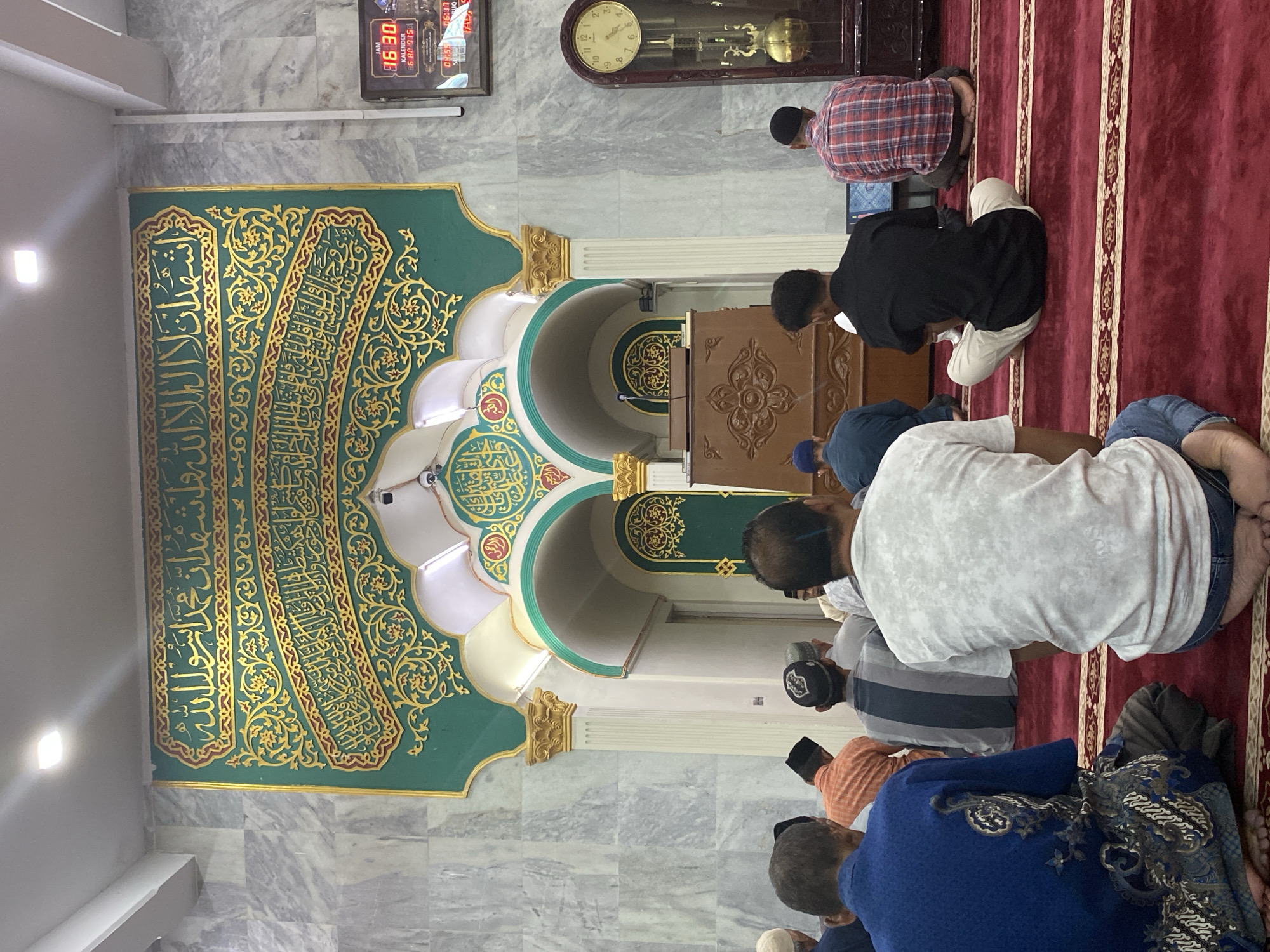 In what appears to be a mosque in Lhokseumawe, Indonesia, several men are lined up on a red carpet, facing an ornate mihrab adorned with Arabic calligraphy in gold on a green background. A wooden minbar sits adjacent to the mihrab. Clocks, including a grandfather clock and a digital clock displaying the time, are mounted on the wall above the men. The floor and walls are covered in gray marble tiles. The men are prostrating in prayer.