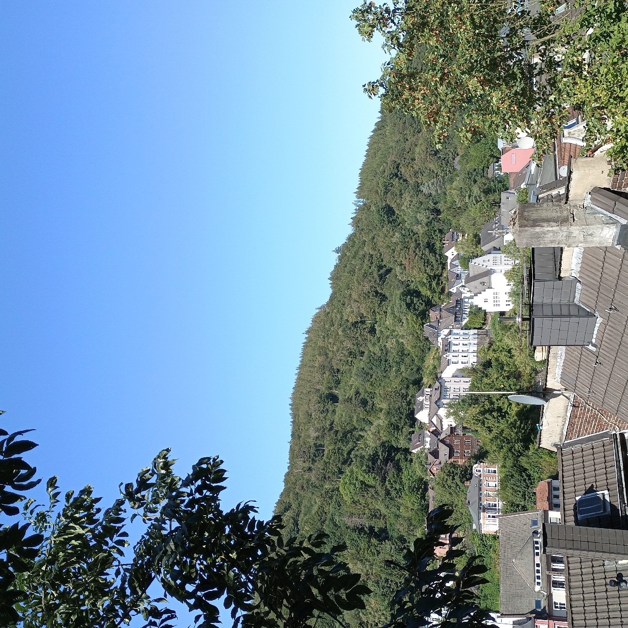 The image shows a town nestled at the foot of a tree-covered hill. The sky is clear and blue. In the foreground, there are several rooftops and chimneys of houses. The buildings in the town are a mix of architectural styles, with many featuring white facades and dark roofs. Some of the buildings are situated on the slope of the hill, suggesting a hilly terrain. The trees on the hill are dense and green, with some patches of lighter green and brown, possibly indicating different tree species or seasonal changes. The overall impression is of a peaceful town surrounded by nature. The vantage point appears to be from a higher elevation, possibly another building or a hill overlooking the town. This scene is consistent with the town of Altena, Germany, which is known for its hilly landscape and traditional architecture.