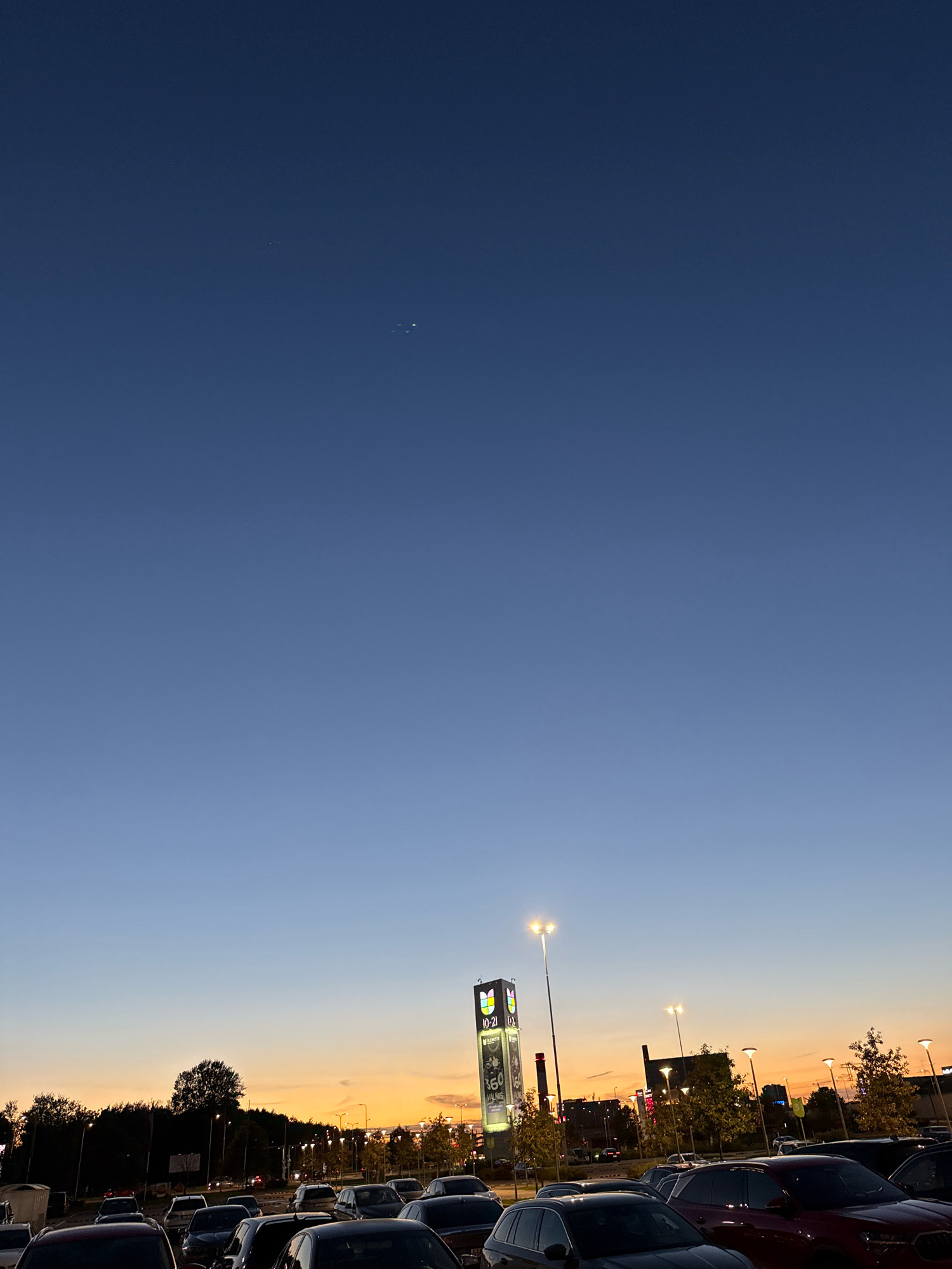 The image depicts a twilight scene in a parking lot, likely in Tallinn, Estonia, given the visible signage with "10-21" and "ELMATE," which are common in Estonian retail establishments. The sky transitions from a deep blue at the top to a soft orange glow near the horizon, indicating sunset or dusk. Several cars are parked in the lot, and streetlights are illuminated, casting a warm light. In the background, there's a tall, illuminated sign structure and other buildings. Trees are visible along the periphery of the parking lot. The overall atmosphere is calm and urban, captured during the transition from day to night.