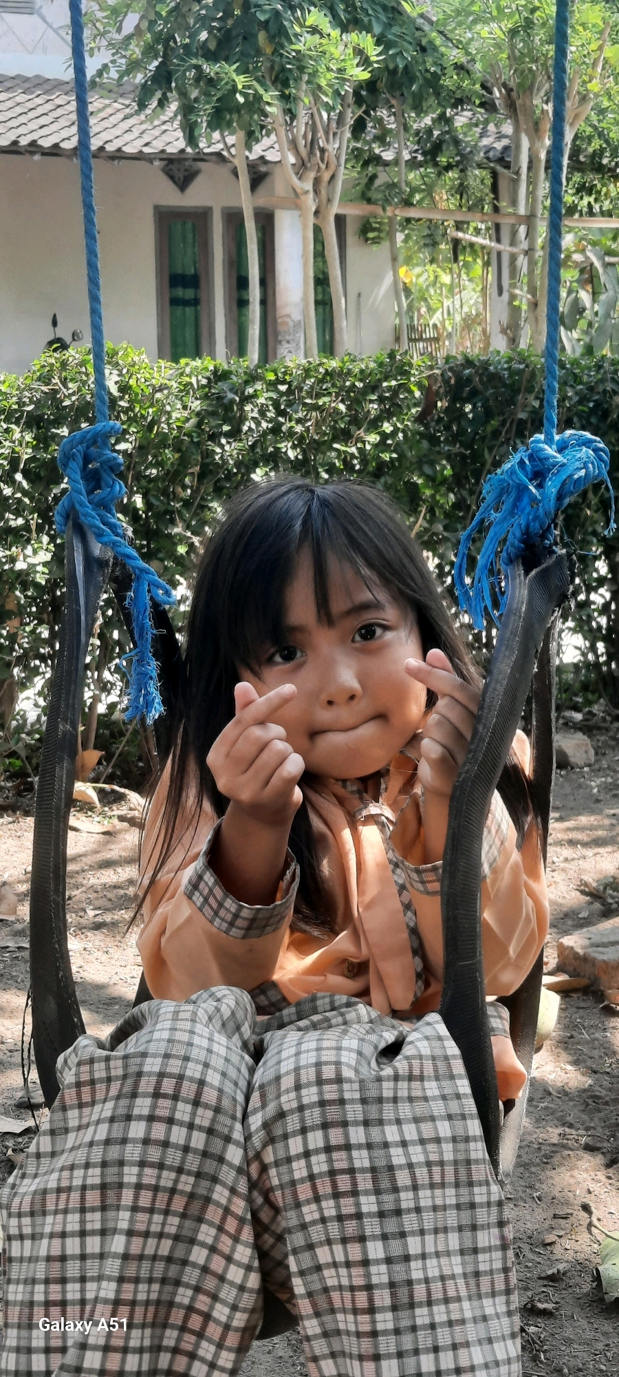 A young girl with long, dark hair is sitting on a swing. She is wearing a light orange shirt with a plaid collar and cuffs, and her legs are covered by a plaid blanket. She is making a heart shape with her fingers on both hands. The swing is made of black tire material and is suspended by blue ropes. Behind her, there is a hedge, trees, and a building with a tiled roof, suggestive of a residential area in Ploso, Indonesia. The ground is covered in dirt and scattered leaves.