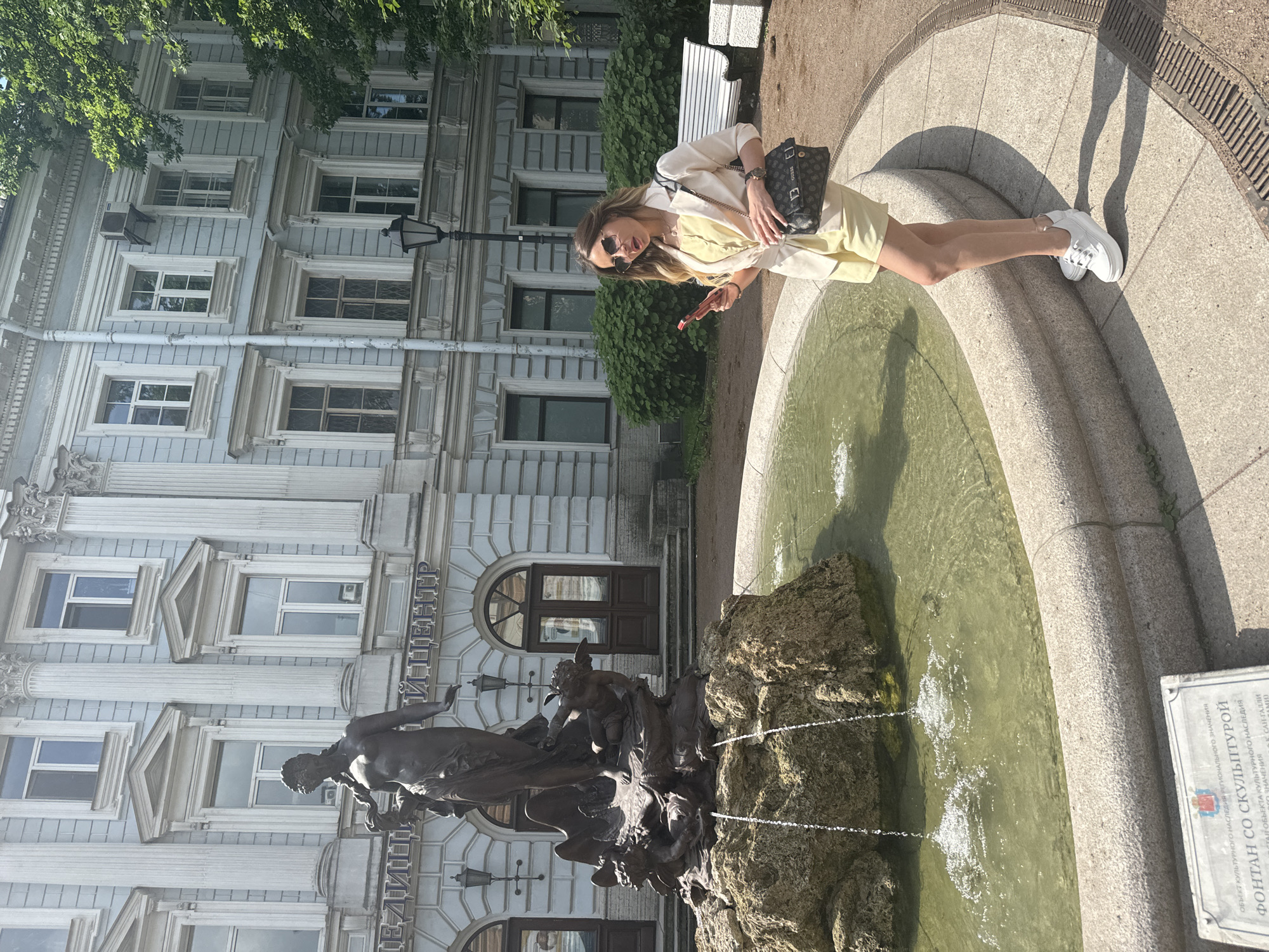 In Saint Petersburg, Russia, a young woman with blonde hair, wearing sunglasses, a light yellow blazer over a matching yellow top and shorts, and white sneakers, stands next to a stone fountain. She is holding a red mobile phone in her right hand and a black handbag with a beige strap hangs from her left shoulder. The fountain features a bronze sculpture of a female figure with a braid, standing on a rock formation with water cascading down. Behind the fountain, an ornate building with classical architecture, featuring columns and arched windows, is visible. Lush green trees and bushes frame the scene, and a white bench is situated in the background. A plaque with Russian text is placed in front of the fountain. The overall setting appears to be a sunny day in an urban park or courtyard.