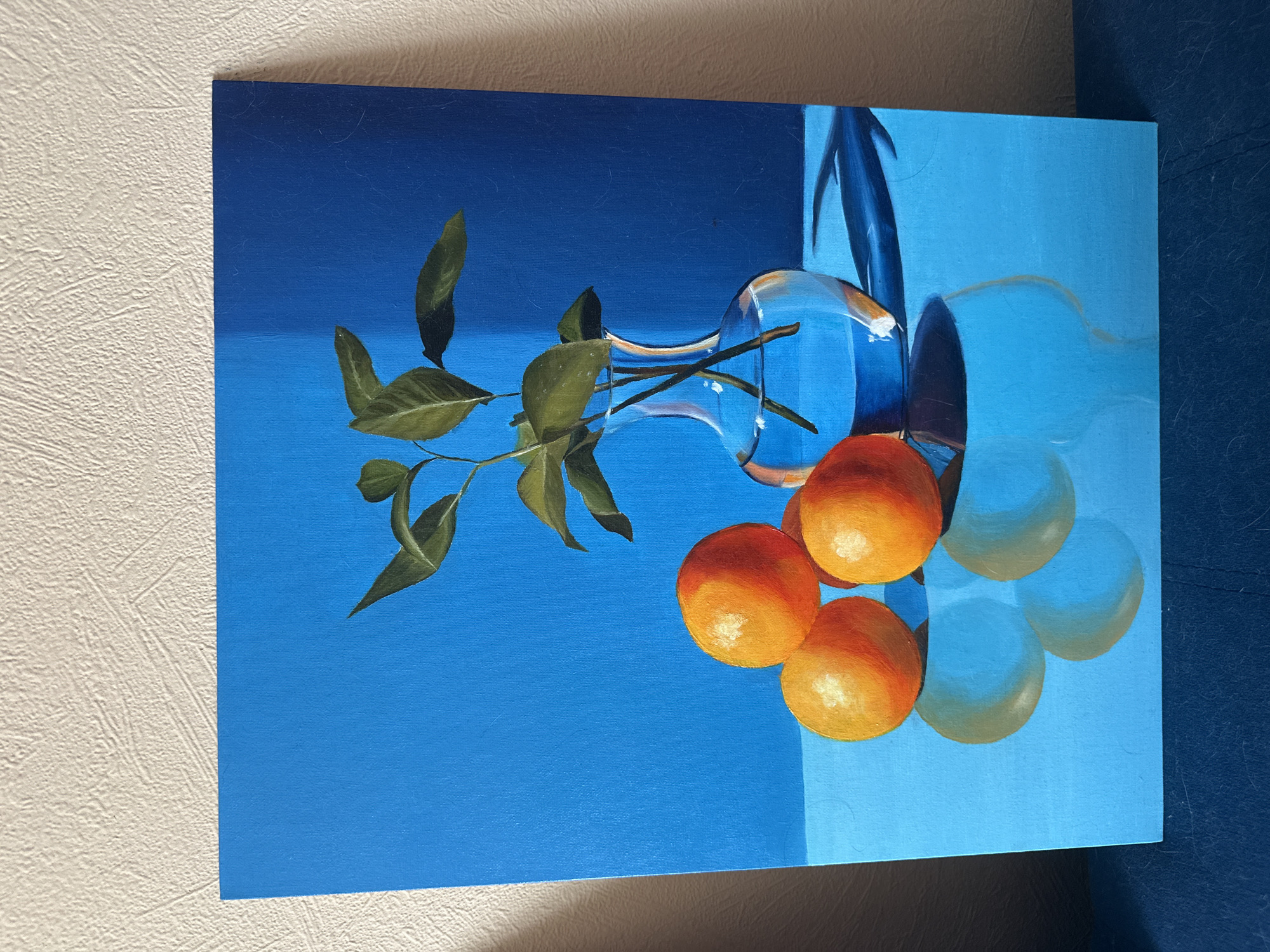 The image shows a still life painting. Three oranges are depicted in the lower right of the canvas. Above the oranges is a clear vase lying on its side with a few green branches inside. The background is divided into two shades of blue, with a lighter blue on the right. The painting is displayed indoors, leaning against a textured white wall on the left and a dark blue wall on the right. No people are visible. The location in the city of Krasnoyarsk, Russia is not discernible from the image.