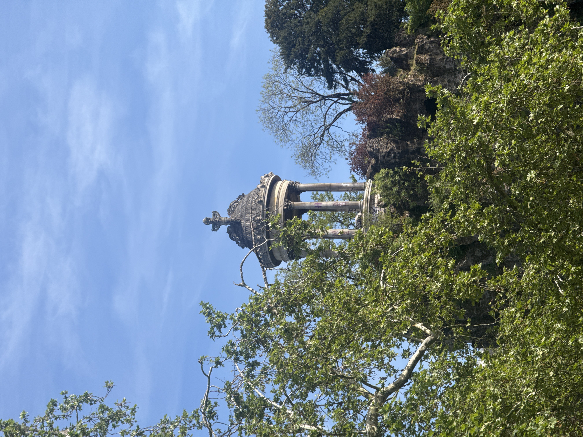 This image, taken in Paris, France, shows a bottom-up perspective of an elaborate stone structure, possibly a monument or temple, with Corinthian columns. The structure sits atop a rocky, elevated area with various trees and foliage surrounding it. The sky is blue with scattered clouds. The image appears to be taken from a park or wooded area, with tree branches and leaves framing the bottom and right of the shot. The monument's details are visible, including its ornamented top and the graffiti on the columns.