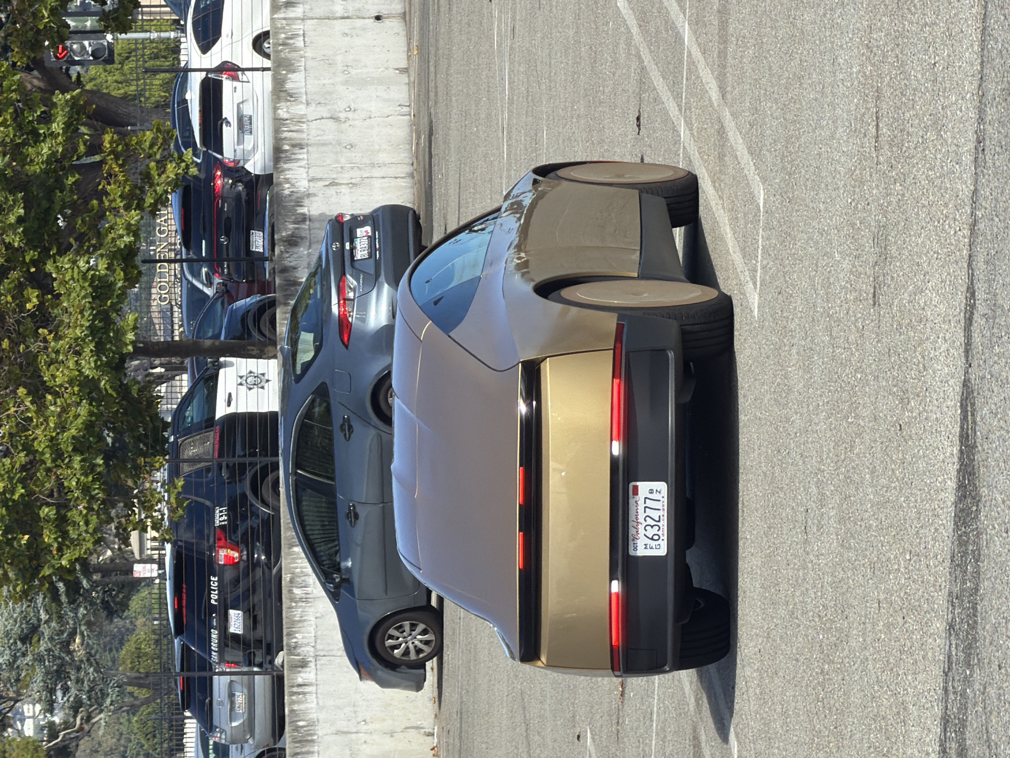 The image shows a collection of parked vehicles on a paved surface, possibly a parking lot or street, with a concrete barrier and a chain-link fence in the background. A large tree with green foliage is on the left side of the frame. The vehicles include several police cars, one of which has "SAN BRUNO POLICE" clearly visible on the back. Another police car has "9-1-1" on its rear. There are also civilian vehicles, including a dark grey sedan and a gold-colored coupe-like car with a prominent license plate reading "OCT California 63277 B".
The scene appears to be outdoors during the daytime, with shadows suggesting bright sunlight. The overall impression is of a static scene, with vehicles parked and no apparent activity. The environment seems urban or suburban, with the presence of official vehicles indicating a public service area or a location where these vehicles are based. The visible text on the police cars and license plates provides specific location and identification details. The image is oriented vertically, with the text appearing sideways.
