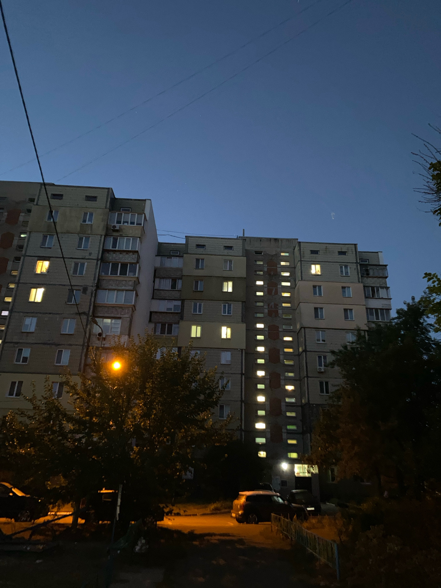 An exterior, eye-level, medium shot captures a tall, multi-story apartment building against a dark twilight sky in an urban setting. The building's facade is constructed of light-colored blocks and features numerous windows, many of which are illuminated with a warm yellow glow, indicating occupied apartments. On the left side of the building, a diagonal black cable stretches across the frame. In the foreground, a street lamp emits a bright orange light, casting illumination on the surrounding trees and a patch of sidewalk. To the right of the street lamp, dense foliage from trees obscures part of the building and foreground. In front of the building, a parked dark-colored car and what appears to be another vehicle are visible, along with a low fence made of horizontal bars. The ground level is a mix of pavement and overgrown vegetation. The sky is a deep, clear blue transitioning to darker hues towards the horizon, with a few faint stars and possibly a distant object visible. The overall scene depicts a residential urban environment at dusk or early night.