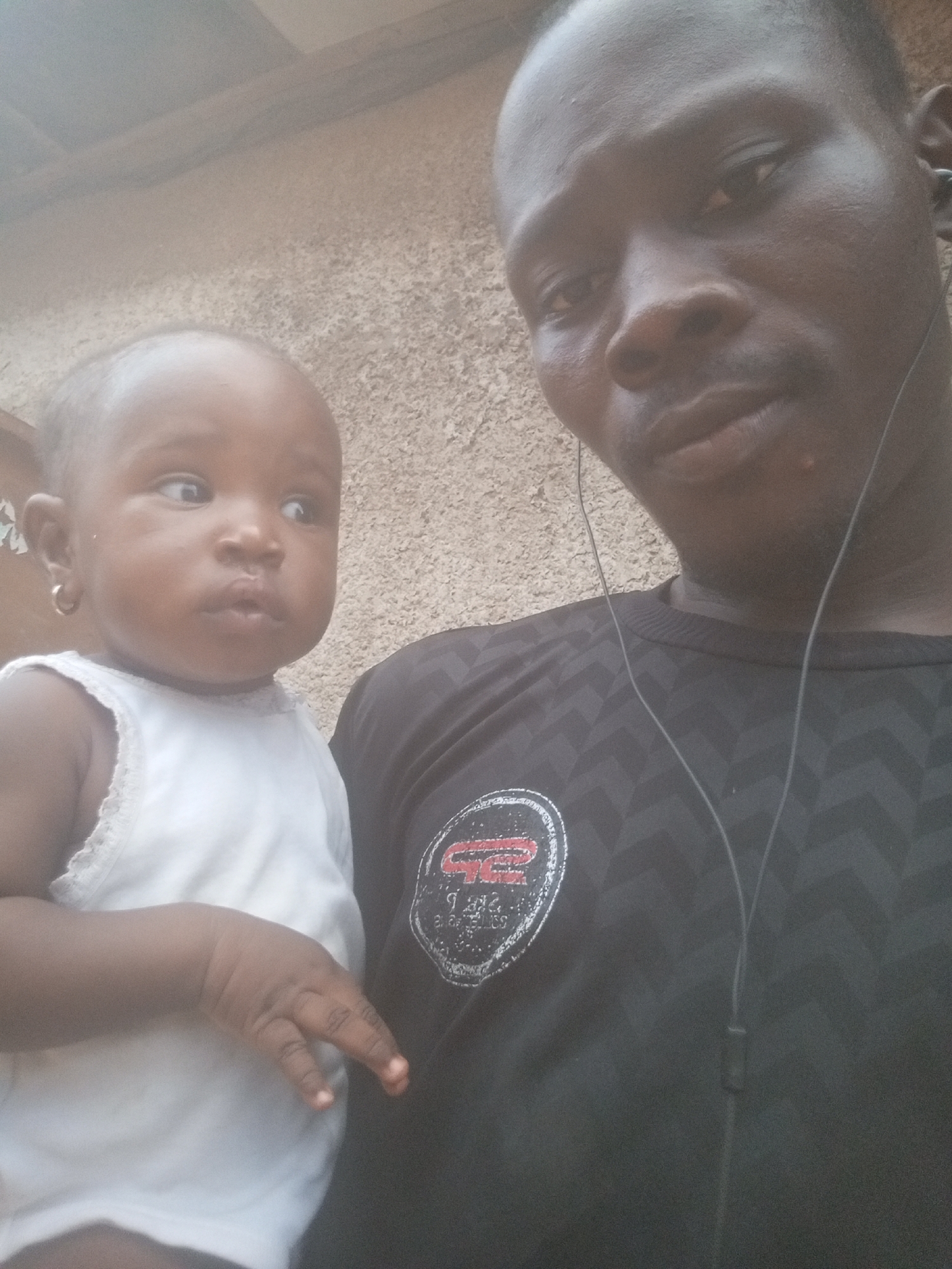 A close-up portrait shows a man holding a baby against a light brown, textured wall. The man, with dark skin and short hair, occupies the right side of the frame, looking directly at the camera with a neutral expression. He is wearing a black long-sleeved shirt that features a subtle dark geometric pattern, possibly chevrons, and a circular logo on his left chest. Black wired earphones are visible around his neck and shoulders.
On the left, he holds a baby, also with dark skin. The baby wears a white sleeveless top with delicate lace trim and has a small gold hoop earring in her left ear. Her gaze is directed slightly to her right, away from the camera, with a curious or contemplative expression. The man's hand is visible, gently supporting the baby's back.
The circular logo on the man's shirt is predominantly red and white on a black background. Visible text on the logo includes "STR" at the top, "SPORT" in the middle, and "COLLECTION" below it, all enclosed within the circular design.
The lighting is soft and even, suggesting either an indoor setting or a shaded outdoor area, possibly on an overcast day. The image captures a quiet, intimate moment between the two subjects, likely in Narayi, Nigeria.