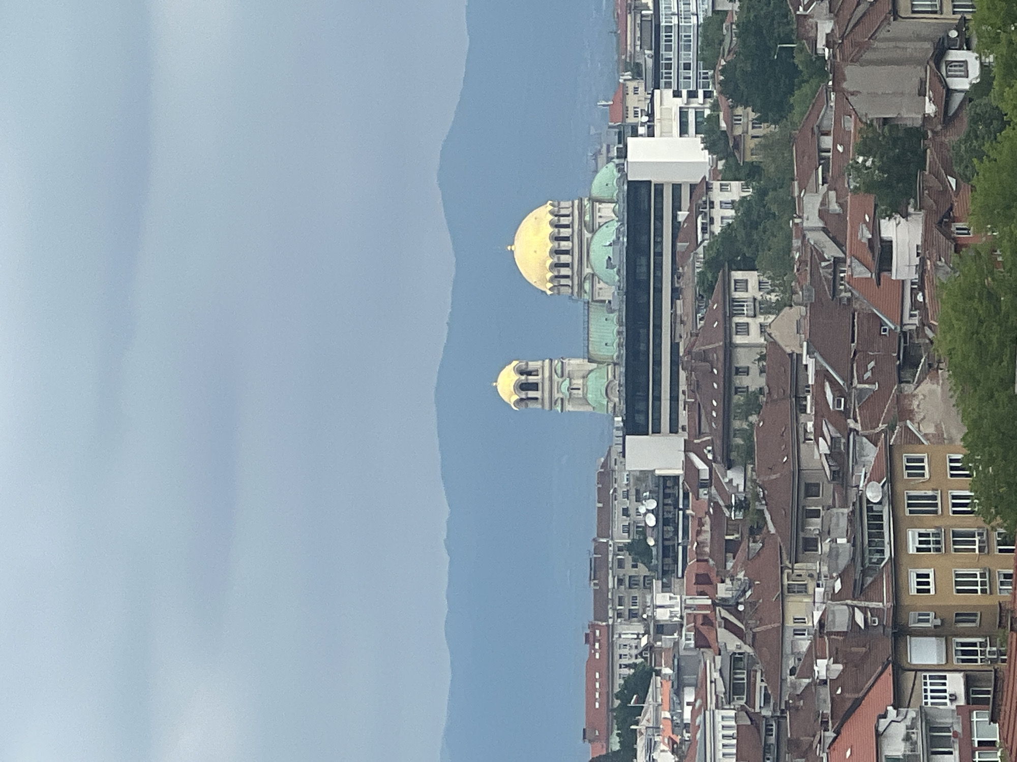 The image depicts an aerial view of a cityscape, likely Sofia, Bulgaria, with a prominent gold-domed Orthodox church visible, appearing to be St. Alexander Nevsky Cathedral. The city's skyline is a mix of older buildings with red tile roofs and modern high-rise structures. Mountains are visible in the background, partially obscured by haze or clouds. No people are readily discernible due to the distance.