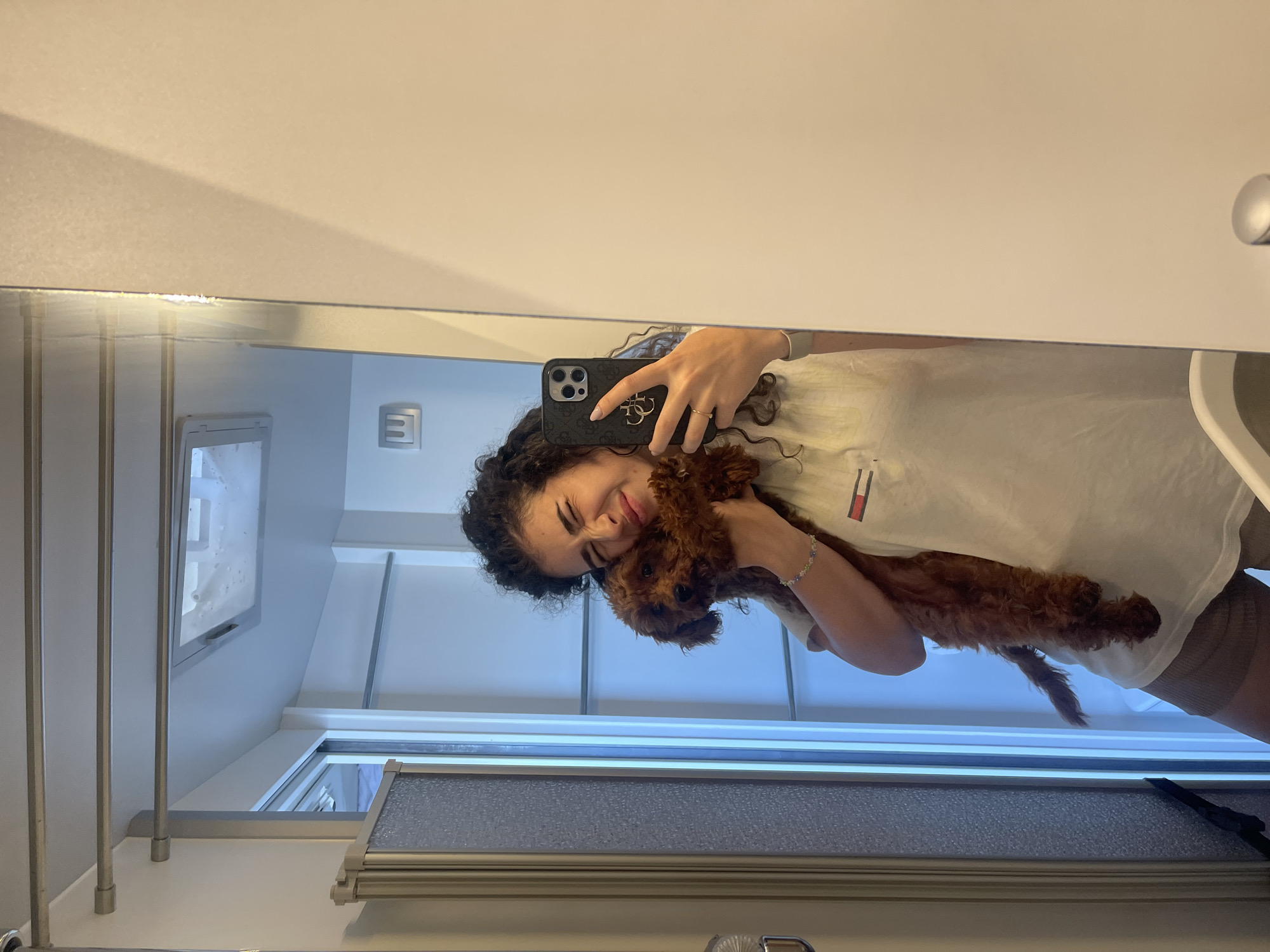 A young woman and a small, reddish-brown curly-haired dog are captured in a mirror selfie in Koprinka, Bulgaria. The woman, with dark curly hair, embraces the dog, holding it close to her face, with her eyes partially closed and a distinct affectionate expression. She is wearing a light-colored t-shirt with a small red, white, and blue flag-like logo (resembling Tommy Hilfiger) on the left chest, and light brown shorts. A multicolored beaded bracelet is on her right wrist, a gold ring on her left ring finger, and a light-colored watch on her left wrist. She holds a dark-colored smartphone, featuring a prominent camera array and a circular pop socket/stand, in her left hand to take the photograph. The dog looks directly towards the camera. The reflection in the mirror reveals an interior space with light-colored walls and ceiling. Visible elements include three horizontal silver rods mounted on the upper left wall, a shower or partition unit with a textured glass or plastic panel and a silver frame to the left of the woman, and a white door frame positioned behind her. A square vent or skylight is present on the ceiling. A rectangular light switch or outlet is reflected on the wall near the woman's head. A silver-capped object, possibly a handle or fixture, is partially visible at the bottom right edge of the mirror. The environment appears clean and compact.