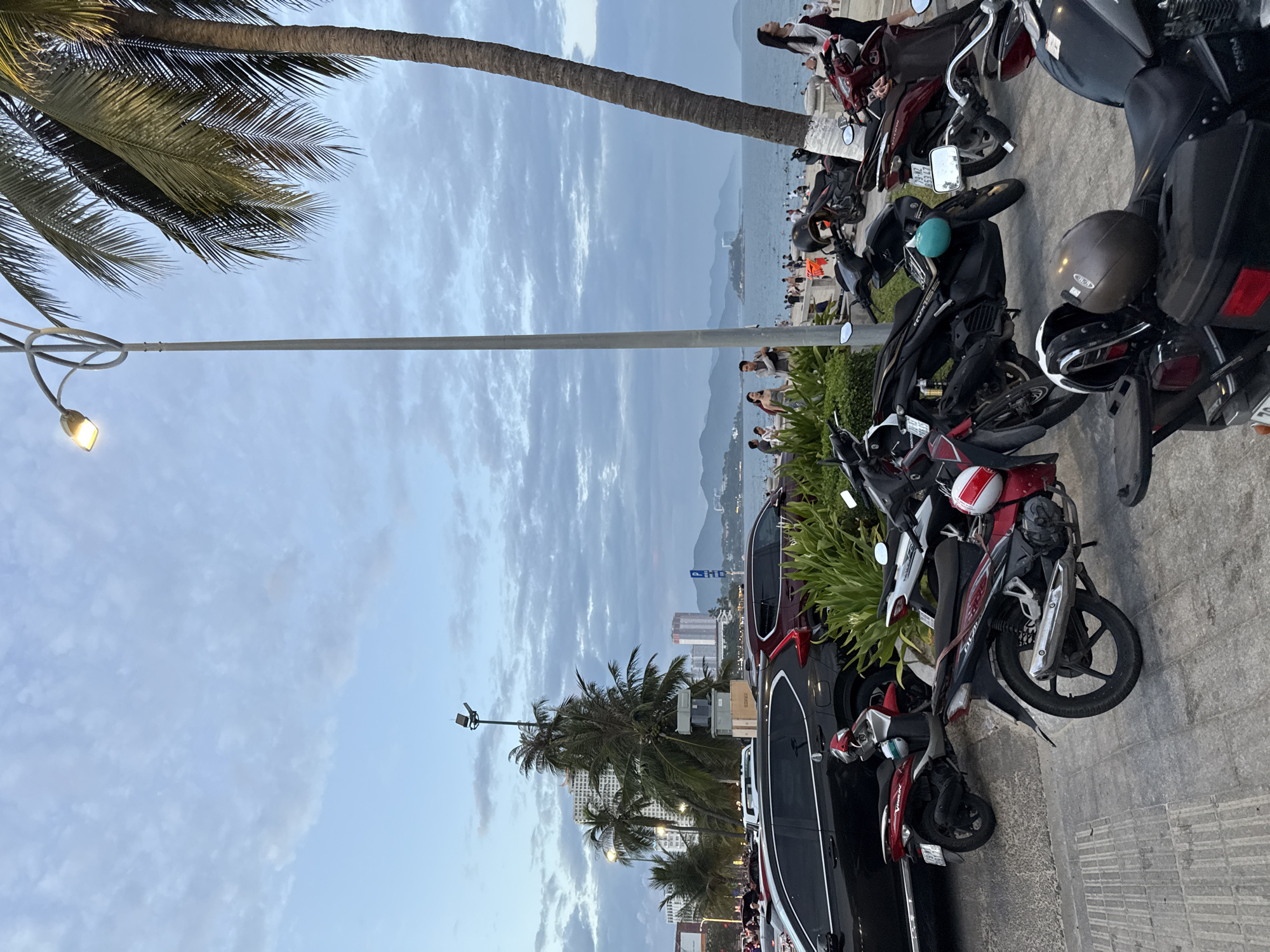 The image depicts a street scene in Nha Trang, Vietnam, near the coastline. Several motorcycles are parked along a paved walkway. In the background, a beach is visible with numerous people enjoying the water and sand. Beyond the beach, the sea stretches to the horizon, with distant islands and mountains. Buildings and palm trees line the street, suggesting an urban coastal environment. The sky is overcast with scattered clouds, and a street lamp is prominently featured in the foreground. The overall atmosphere suggests a moderately busy day at a popular tourist destination.