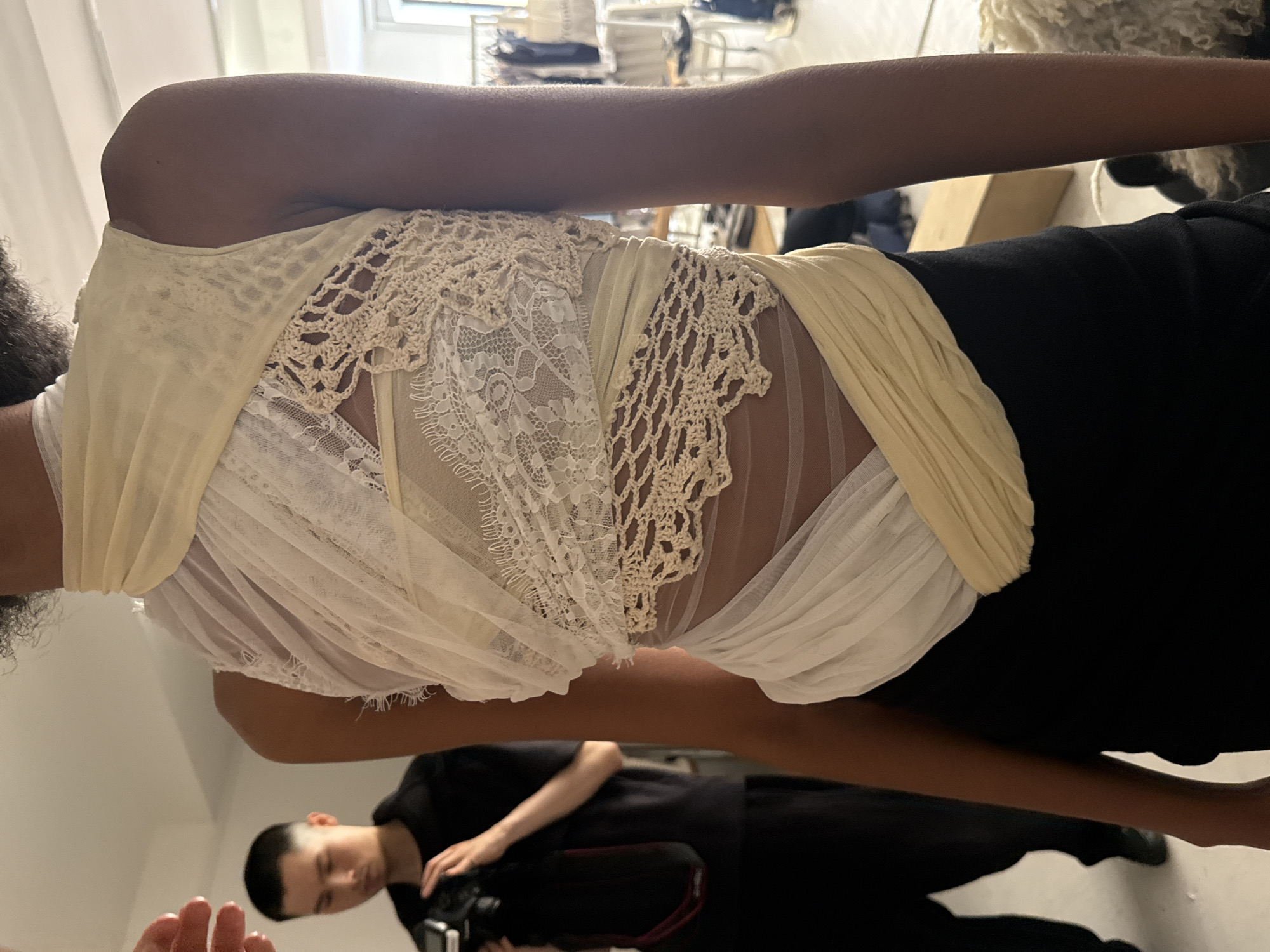 The image depicts a person wearing a white lace and crochet-detailed top with draped detailing, paired with black pants. The person is photographed from a low angle, with another individual, presumably a photographer, visible in the reflection. The environment appears to be a studio or indoor setting with garments hanging in the background. Based on the available information, the location is not discernible as being specifically in London, United Kingdom.