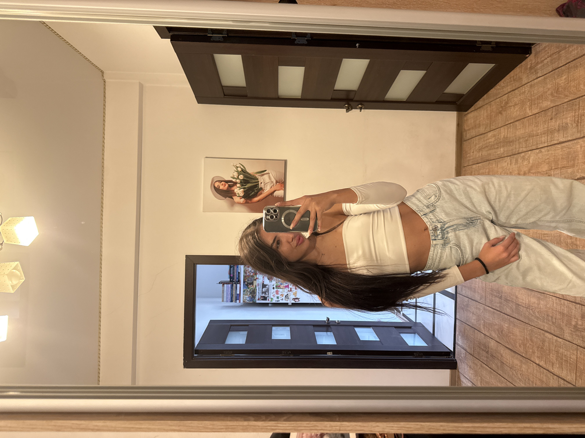 A young woman with long dark hair takes a mirror selfie in a hallway. She is wearing a white long-sleeved crop top and light-wash baggy jeans. She is holding a smartphone in her right hand, angled to capture her reflection. To her left, a doorway is open, revealing a glimpse of another room with shelves filled with books and items. To her right, a dark wood door with frosted glass panels is partially ajar. On the wall behind her, a framed picture depicts a woman in a hat holding a bouquet of tulips. The hallway has wood-paneled flooring and a light-colored wall. The lighting suggests it is daytime, possibly indoors. There is no indication of the city of Chișinău, Moldova in the image.