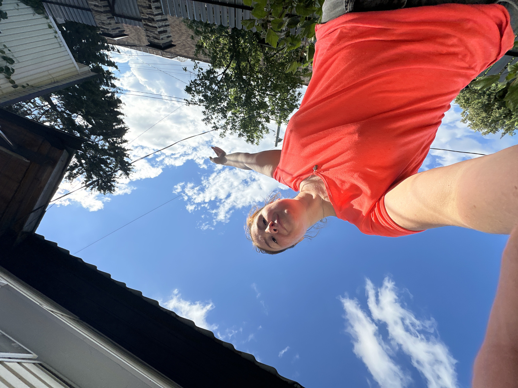 The image shows a low-angle, close-up shot of a young woman with auburn hair wearing an orange t-shirt. She is looking up at the camera with a slight smile, her face tilted. Her right arm is extended upwards, pointing towards the sky. The background is a bright blue sky with scattered white clouds.
The scene appears to be outdoors, with parts of a building and some greenery visible in the upper left and right corners of the image. A dark-colored roofline is prominent in the lower part of the frame. The lighting suggests it is daytime, likely sunny, with the sun casting shadows that are not clearly visible due to the angle.
The overall mood is casual and playful, with the woman's upward gaze and outstretched arm suggesting a moment of joy or engagement with the camera. The image seems to be a selfie taken from a low perspective. No visible text is present. The location context is Nizhny Novgorod, Russia.