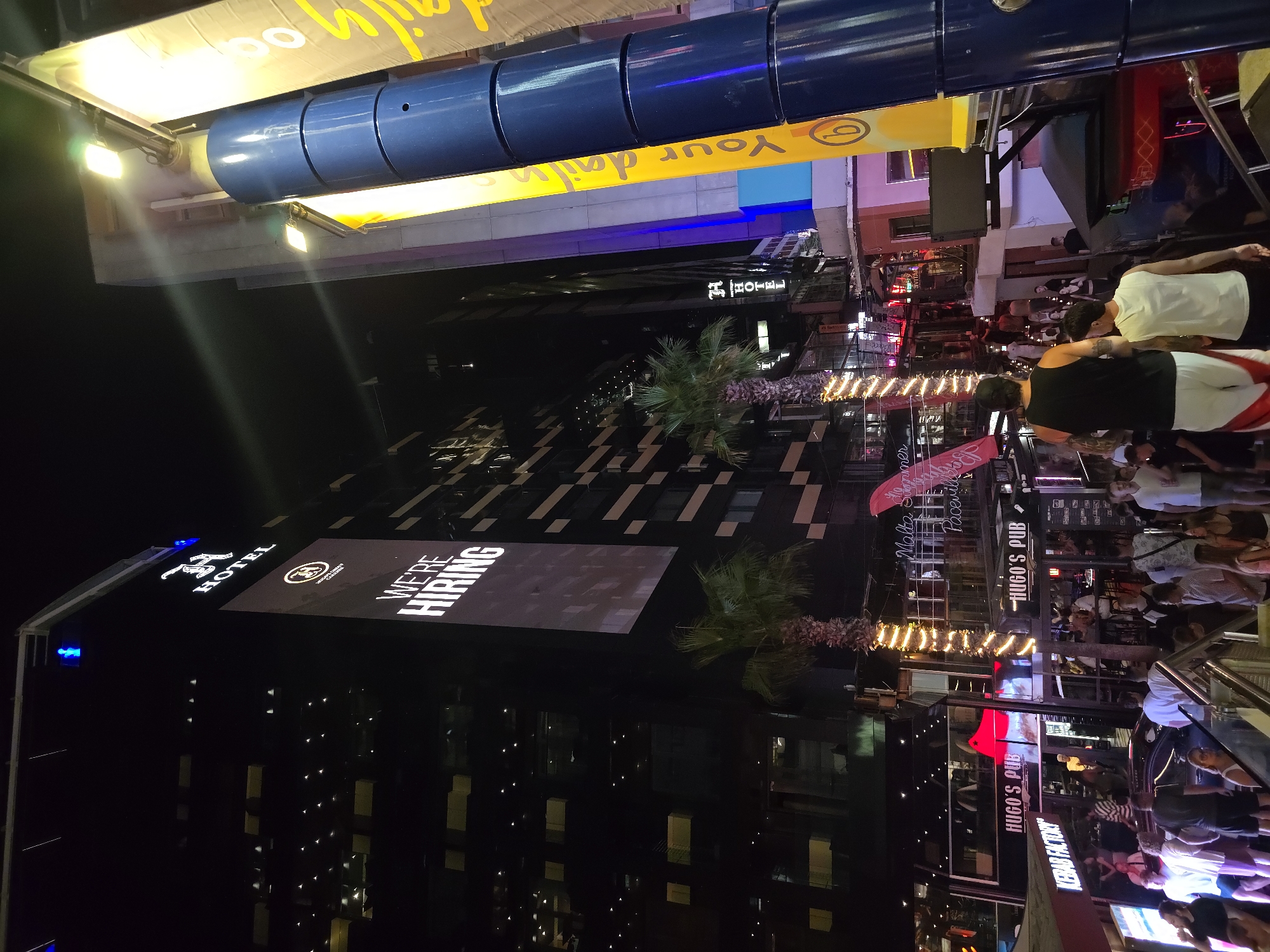 The image depicts a vibrant nighttime scene in Saint Julian's, Malta, likely Paceville, a popular entertainment district. The foreground features a crowd of people on a street or staircase, with a focus on two men walking away from the camera. The architecture is modern and tall, with the prominent "H HOTEL" sign illuminated on a dark, glass-fronted building. A large digital billboard on the hotel displays "WE'RE HIRING" in white text against a dark background. Further into the scene, "Hugo's Pub" is visible with external seating and patrons. Other establishments like "Kebab Factory" can also be identified. The street is lined with palm trees, some decorated with festive lights. Tall, cylindrical blue structures and bright yellow banners with text suggest commercial advertising and street fixtures. The overall atmosphere is lively and bustling, characteristic of a nightlife area with numerous bars and businesses.