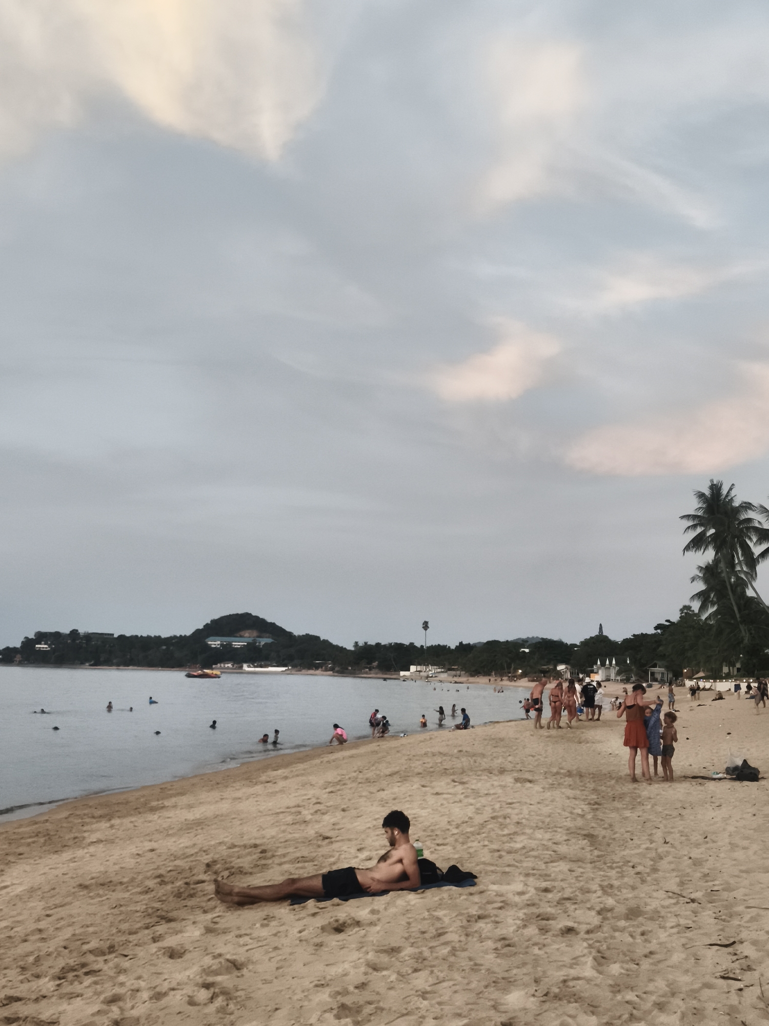 A serene beach scene unfolds on the coast of Ko Samui District, Thailand, under a soft, cloudy sky. The foreground is dominated by a sandy beach with footprints and some scattered debris. A man, wearing black swim trunks, is lying on his side on the sand, facing away from the camera, with his left arm bent and resting near his head. A black towel is placed under him.
Further back, the calm sea laps gently onto the shore. Several people are visible in the water, some wading and others swimming. A yellow inflatable raft is floating on the water's surface.
Along the shoreline, a group of people are gathered. Some are walking, others are standing and talking. A woman in a reddish-orange dress is seen with two children. Further down the beach, a line of palm trees and lush vegetation marks the edge of the land, with some buildings and structures visible in the distance. A small hill or mountain covered in trees is silhouetted against the horizon. The overall impression is of a relaxed, late afternoon on a tropical beach.